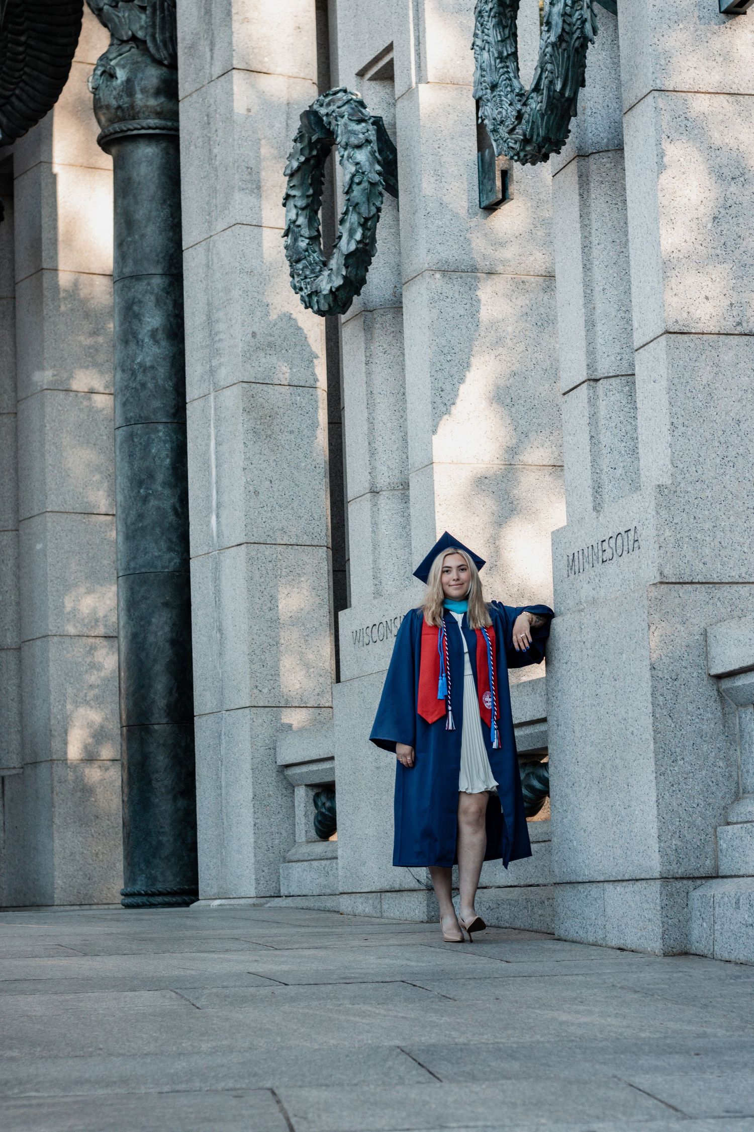 Graduates pose between architectural columns in blue graduation regalia with red accents.