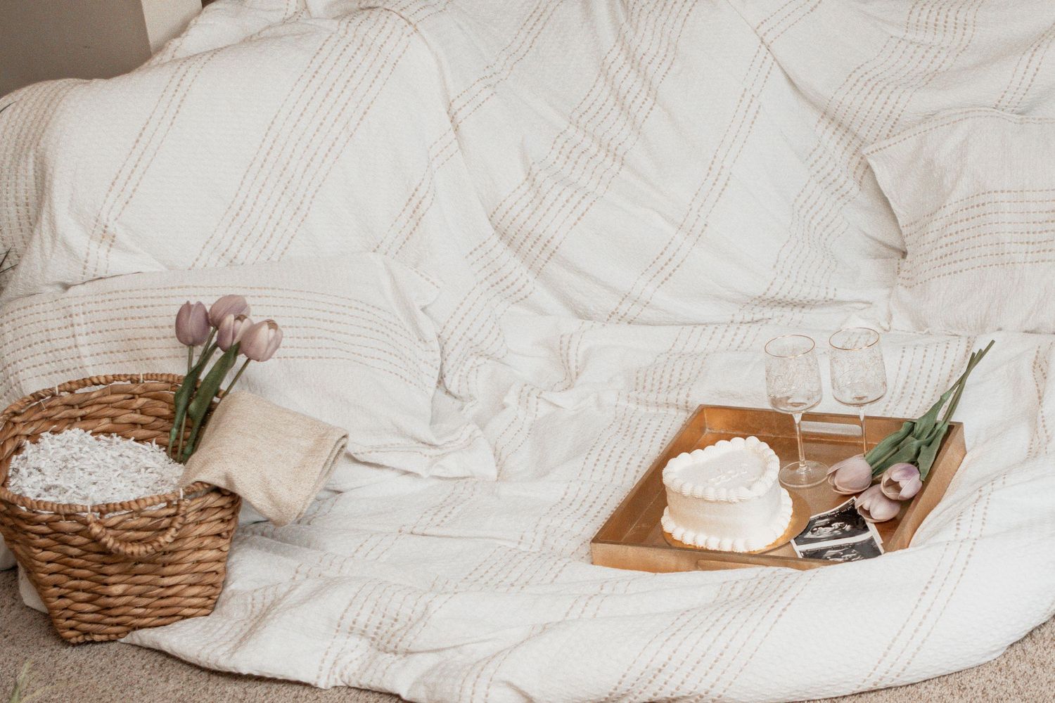 Cozy white bed linens with wicker basket and wooden tray holding flowers and decorative items against neutral background.