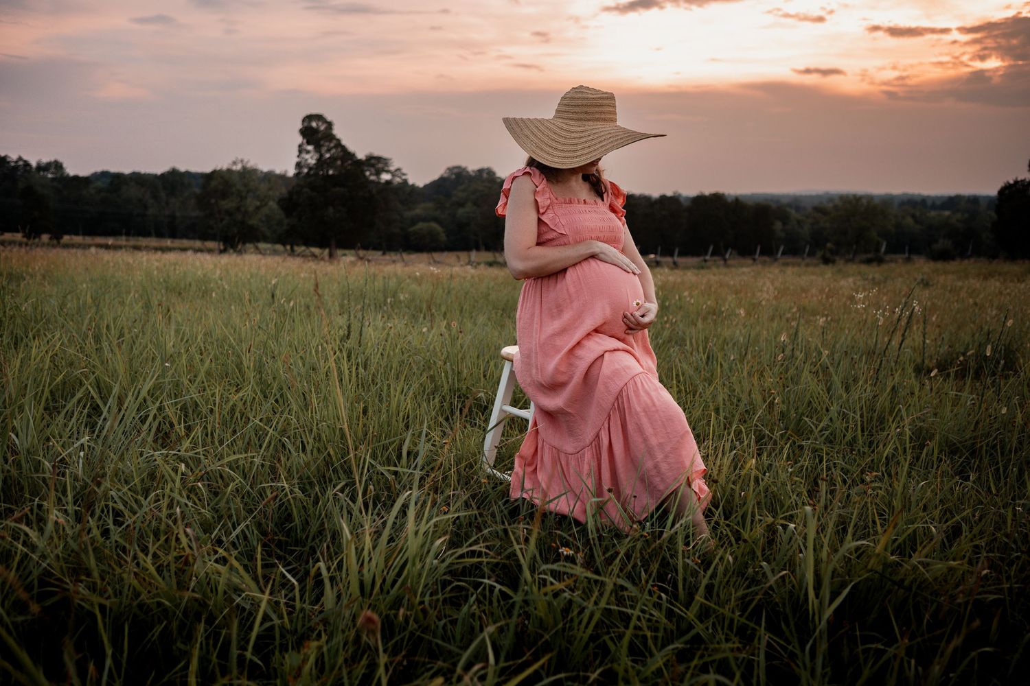 Figure in a pink flowing dress and wide brim hat walks through a grassy meadow at sunset.