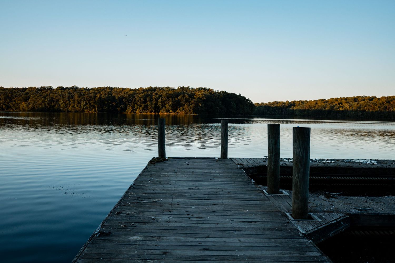 Wooden dock extends over calm lake waters during sunset with trees and shoreline visible in the distance.