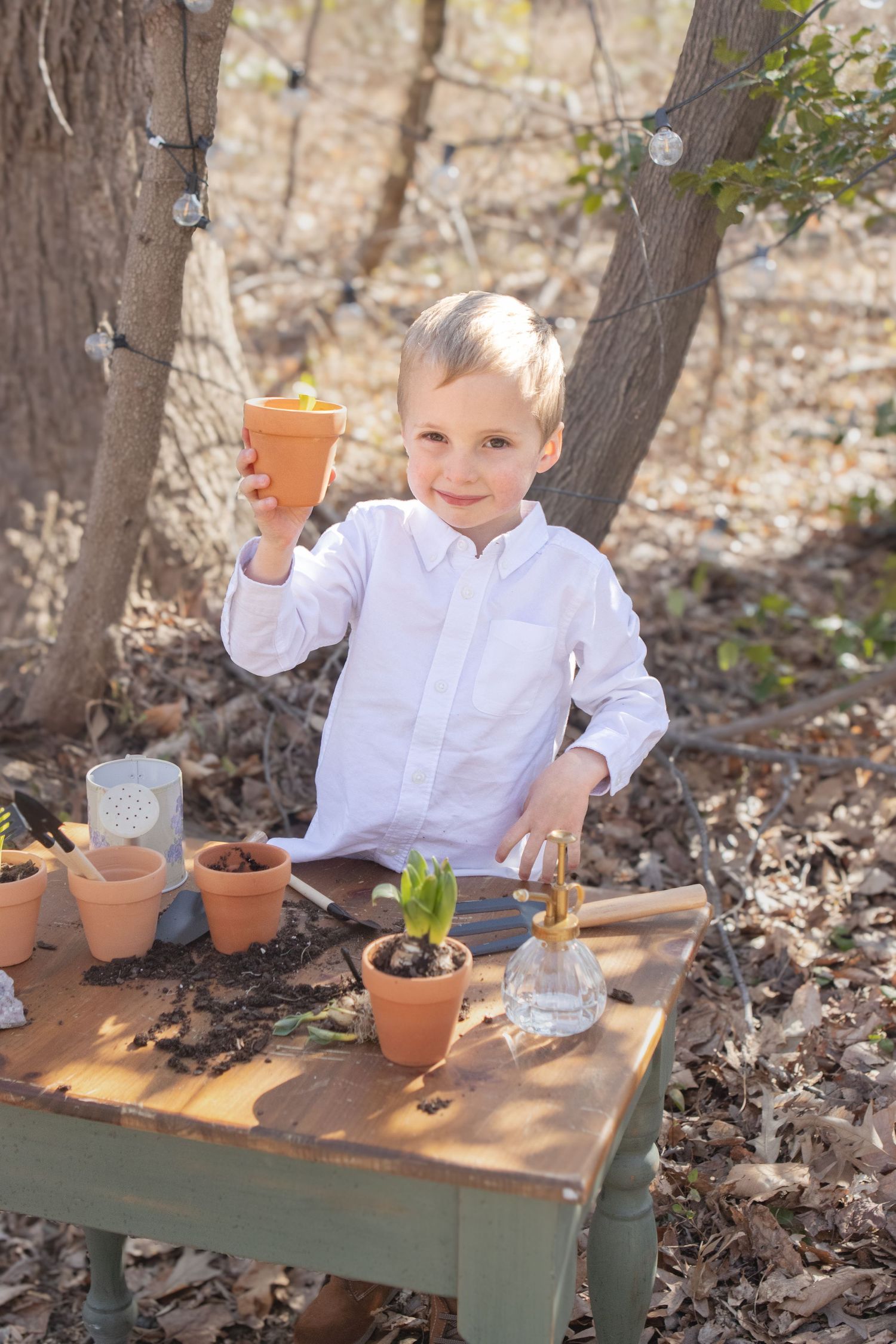 Child in white shirt proudly holding terracotta pot at outdoor gardening table.