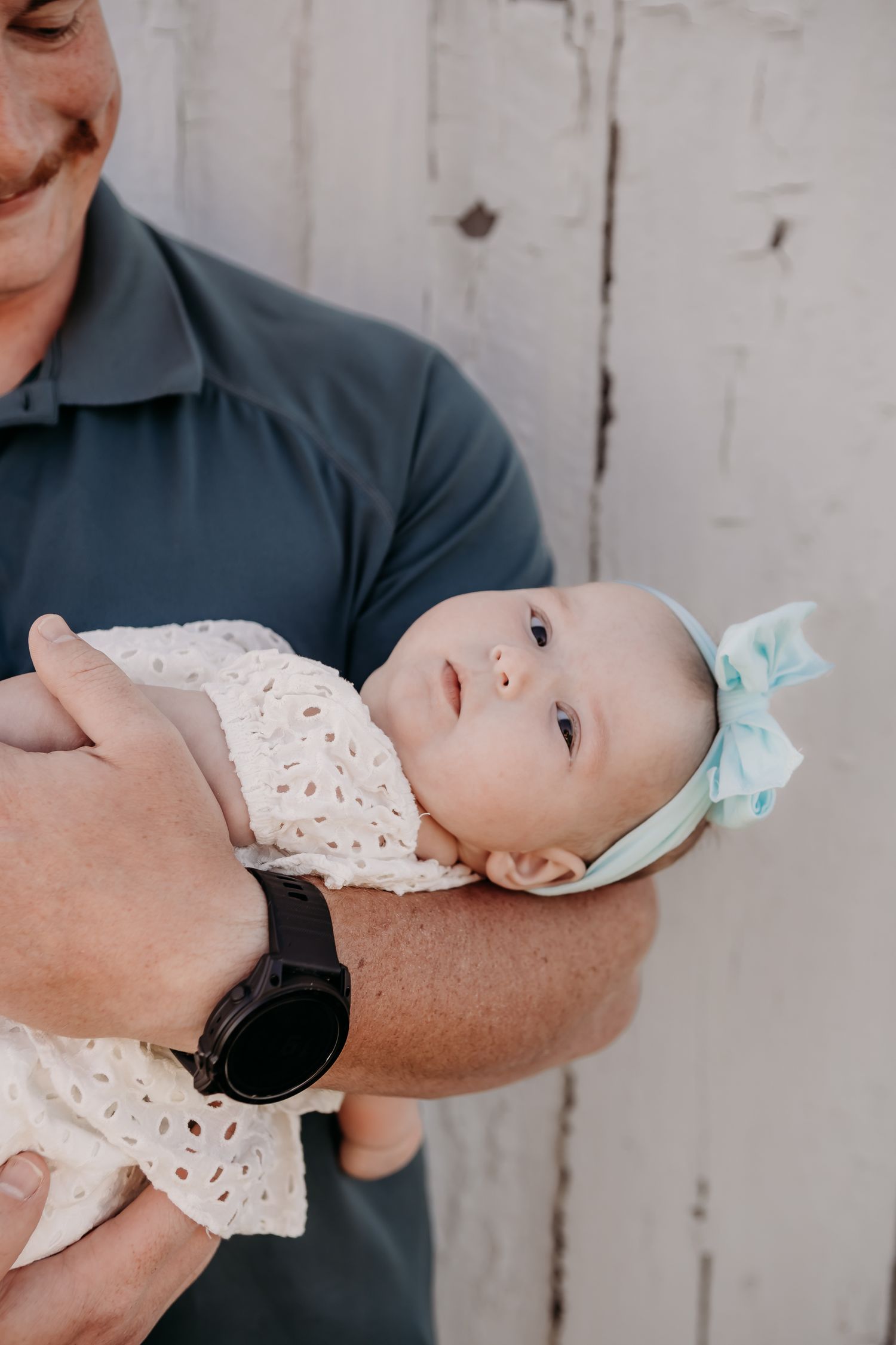 A baby wearing a light colored dress and blue bow headband is being held in someone's arms.