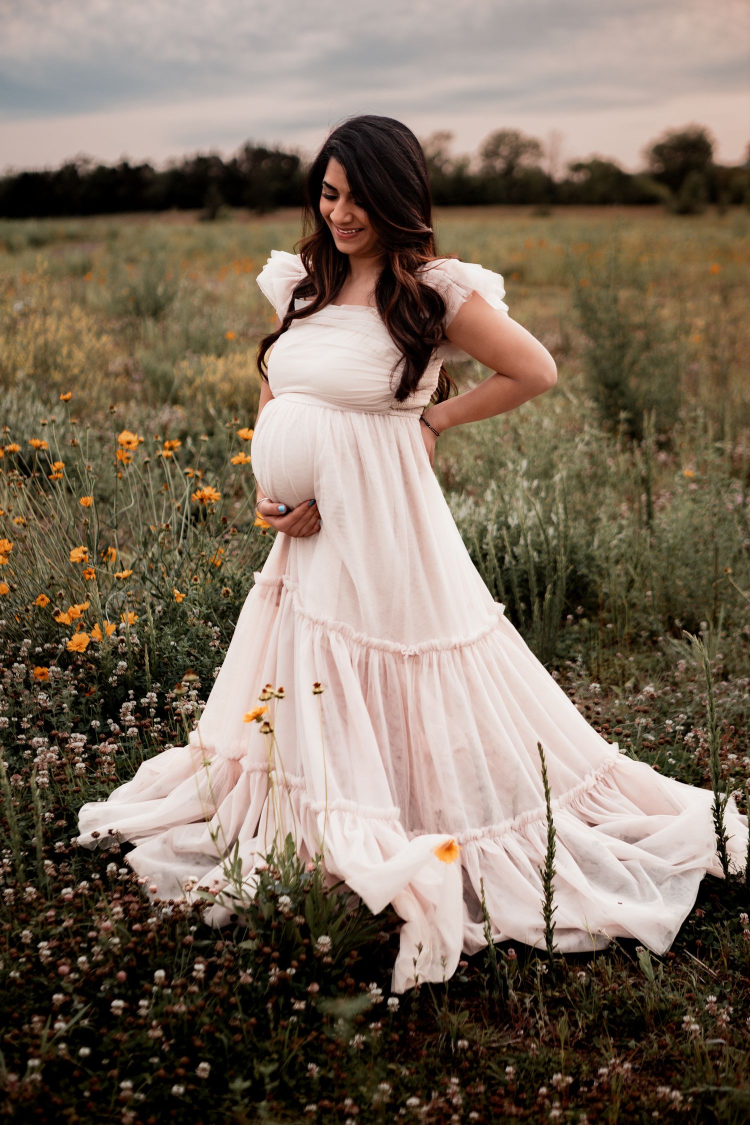 Pregnant woman in flowing white maxi dress stands in wildflower field at sunset with orange blooms and tall grass.