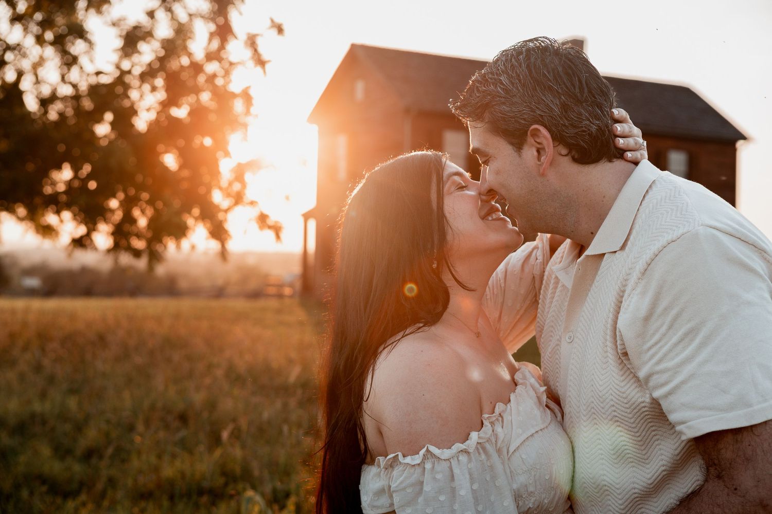 A romantic couple embraces in a golden sunset with a rustic barn in the background.