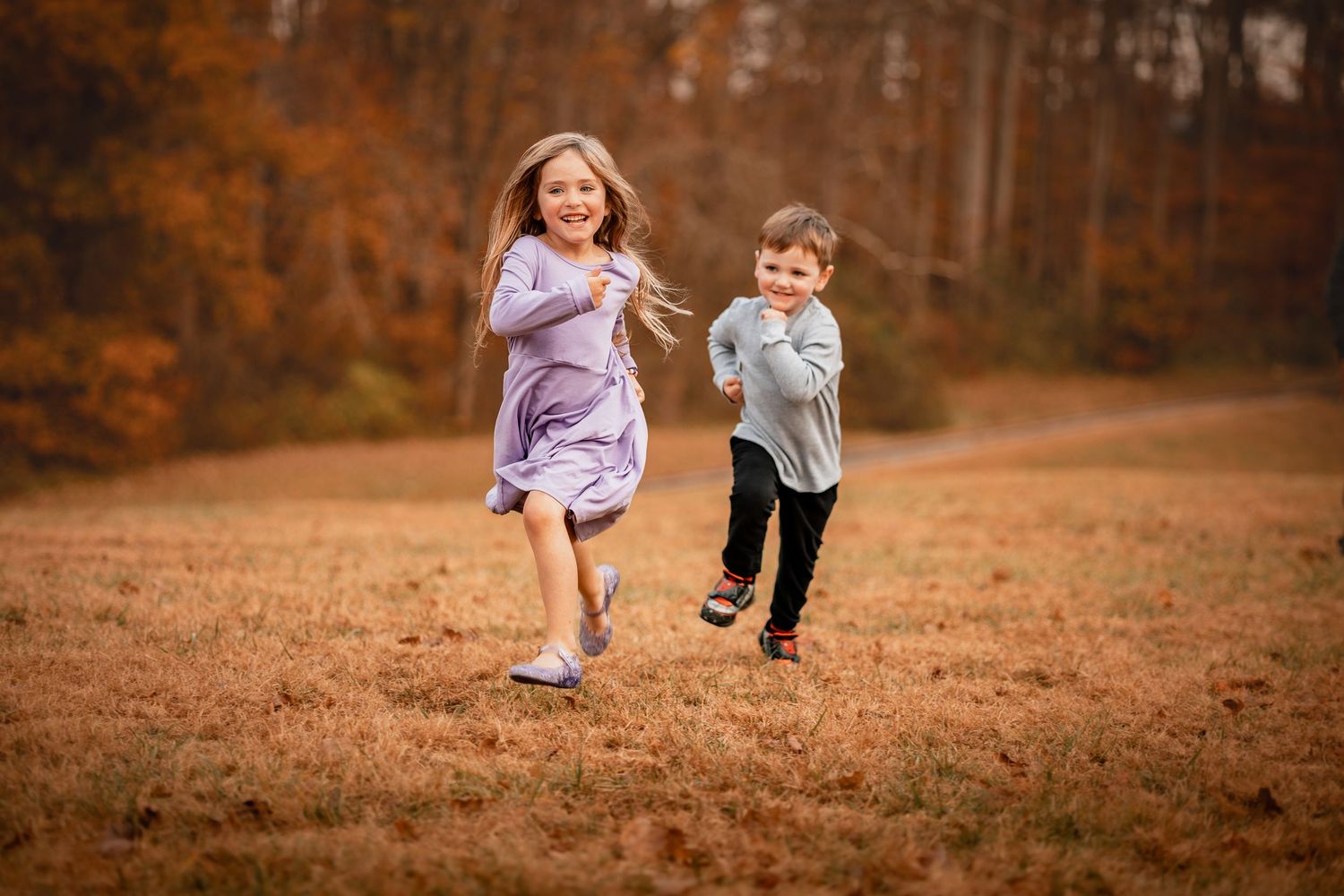 Children chase each other playfully through a brown autumn field with trees in the background.