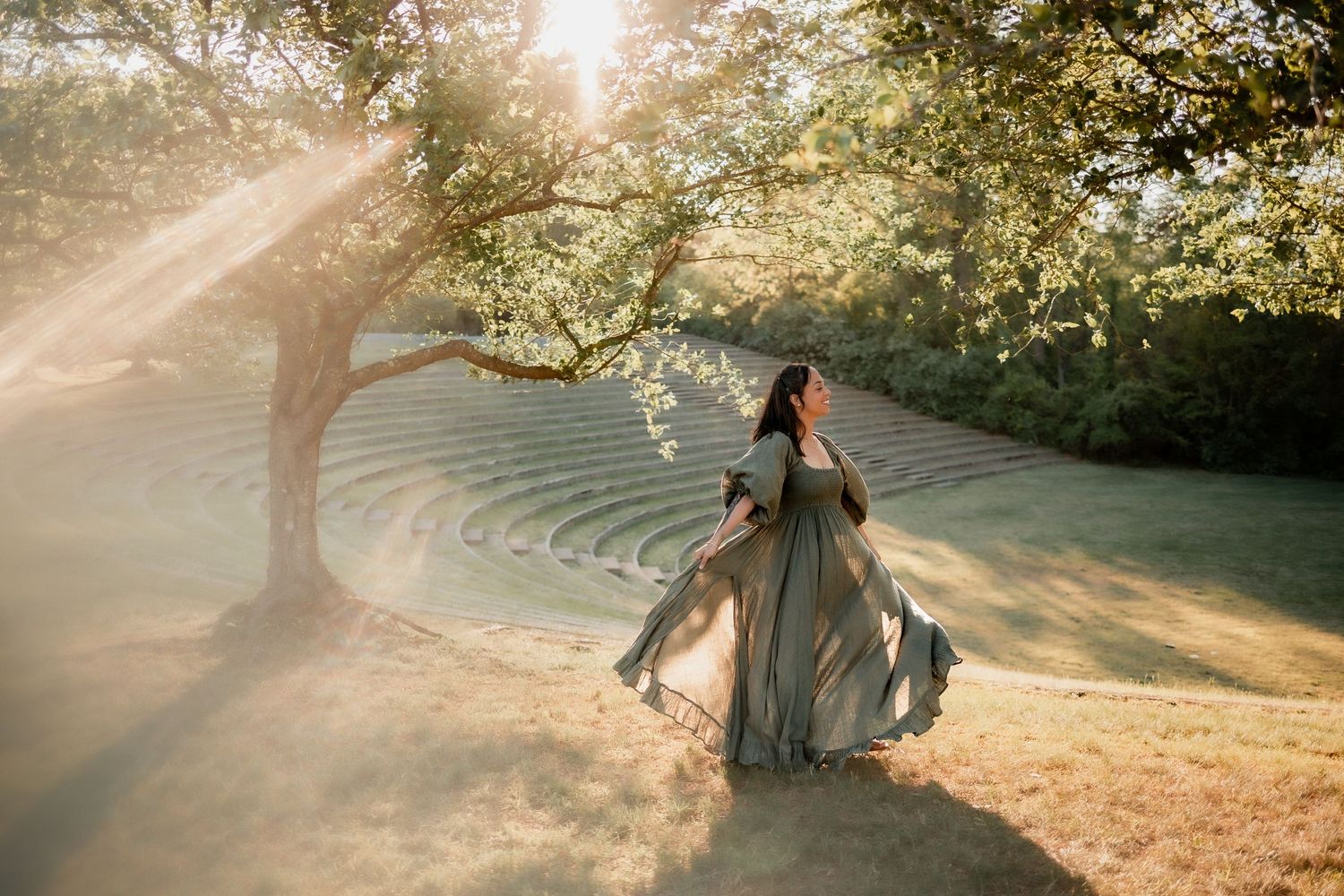 Woman in flowing gray gown dancing under sunlit tree near outdoor amphitheater.