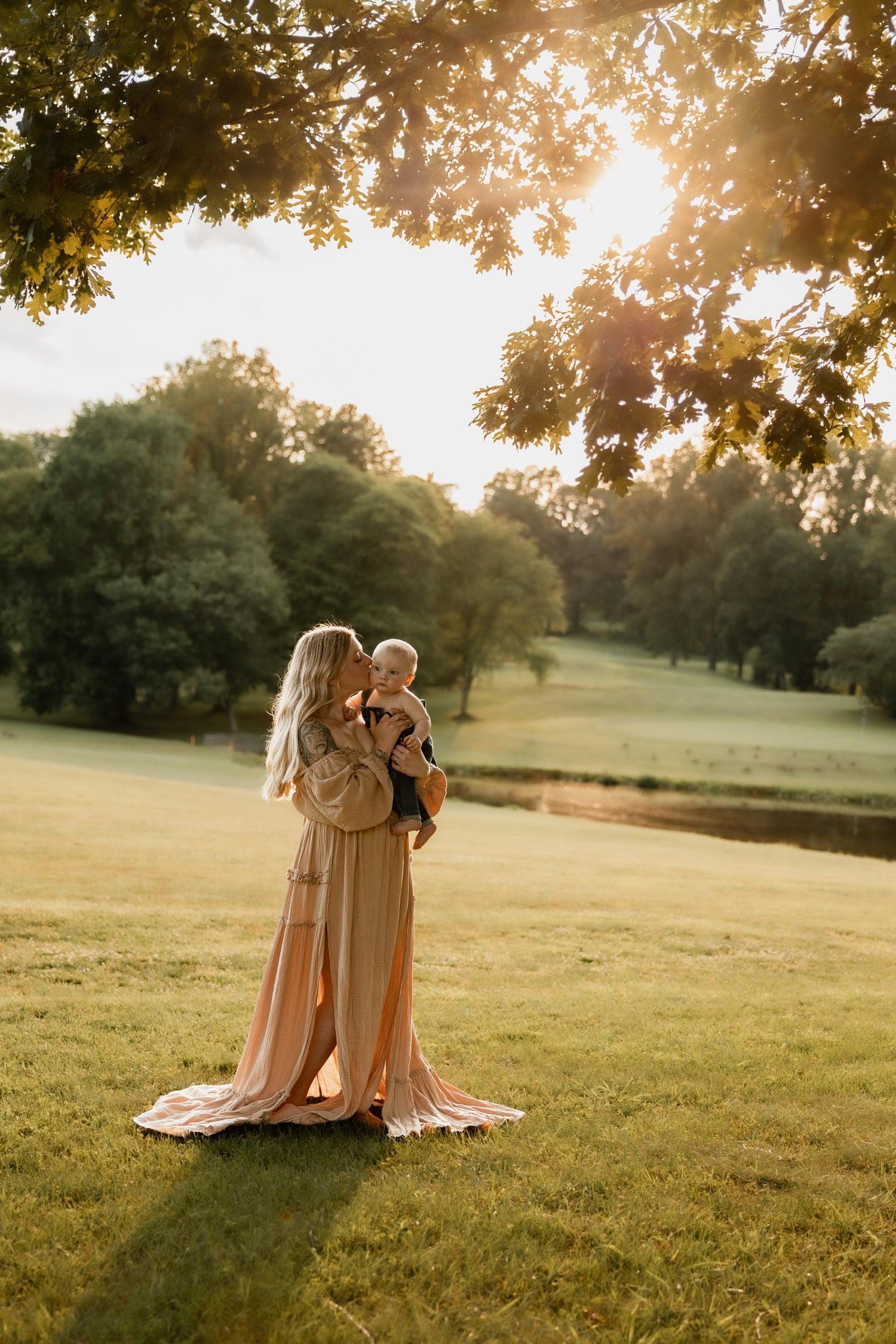 Mother in pink gown kissing baby at golden hour in lush green park under oak tree.