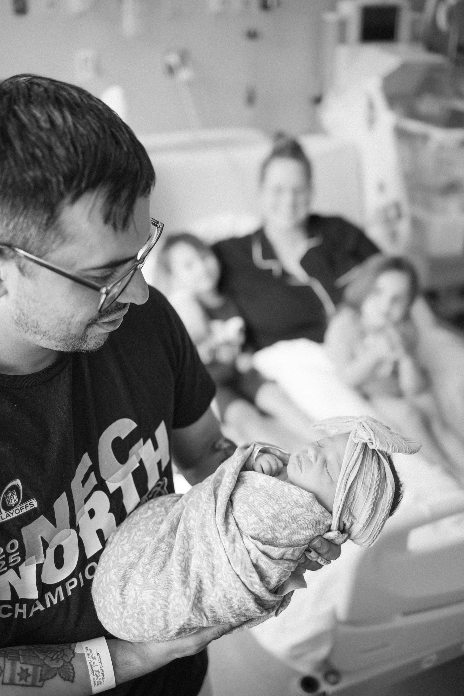 Father holds swaddled newborn in hospital room while mother rests in bed with older child in black and white photo.