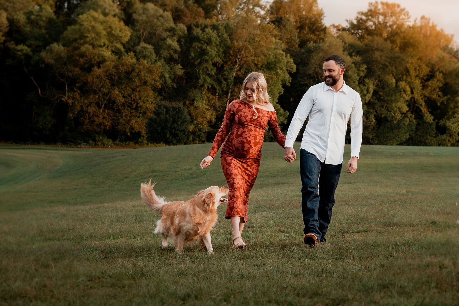 A couple walks with their golden retriever through an autumn field at sunset as warm golden light illuminates the scene.