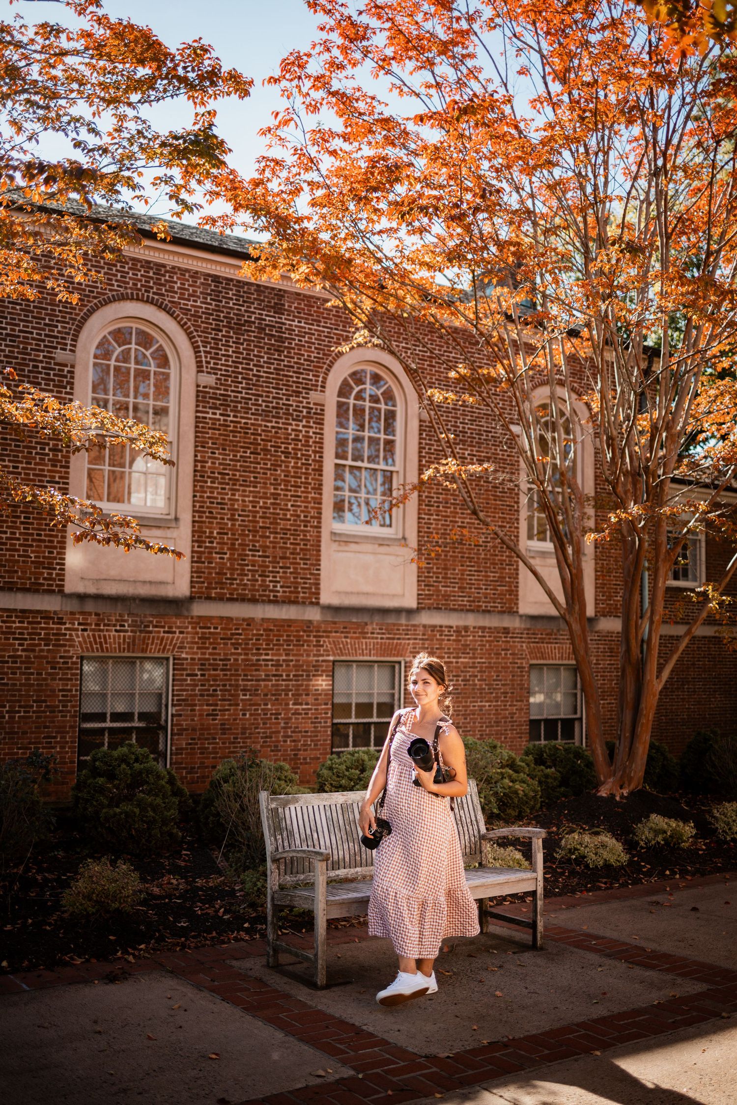 Autumn trees with vibrant orange leaves frame a brick building with white windows and a bench in the courtyard.