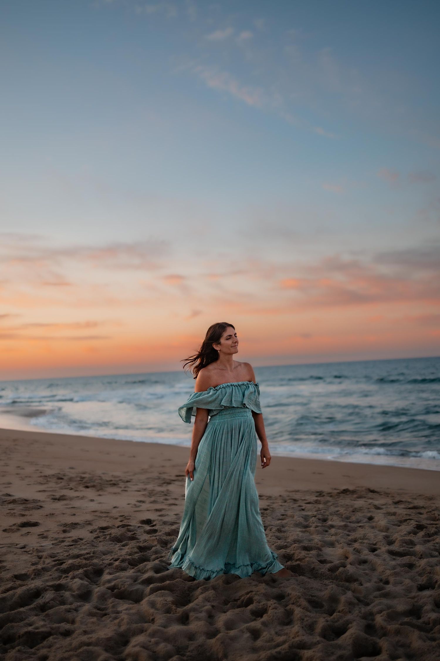 Woman in teal off-shoulder maxi dress standing on sandy beach at sunset with ocean waves behind her.