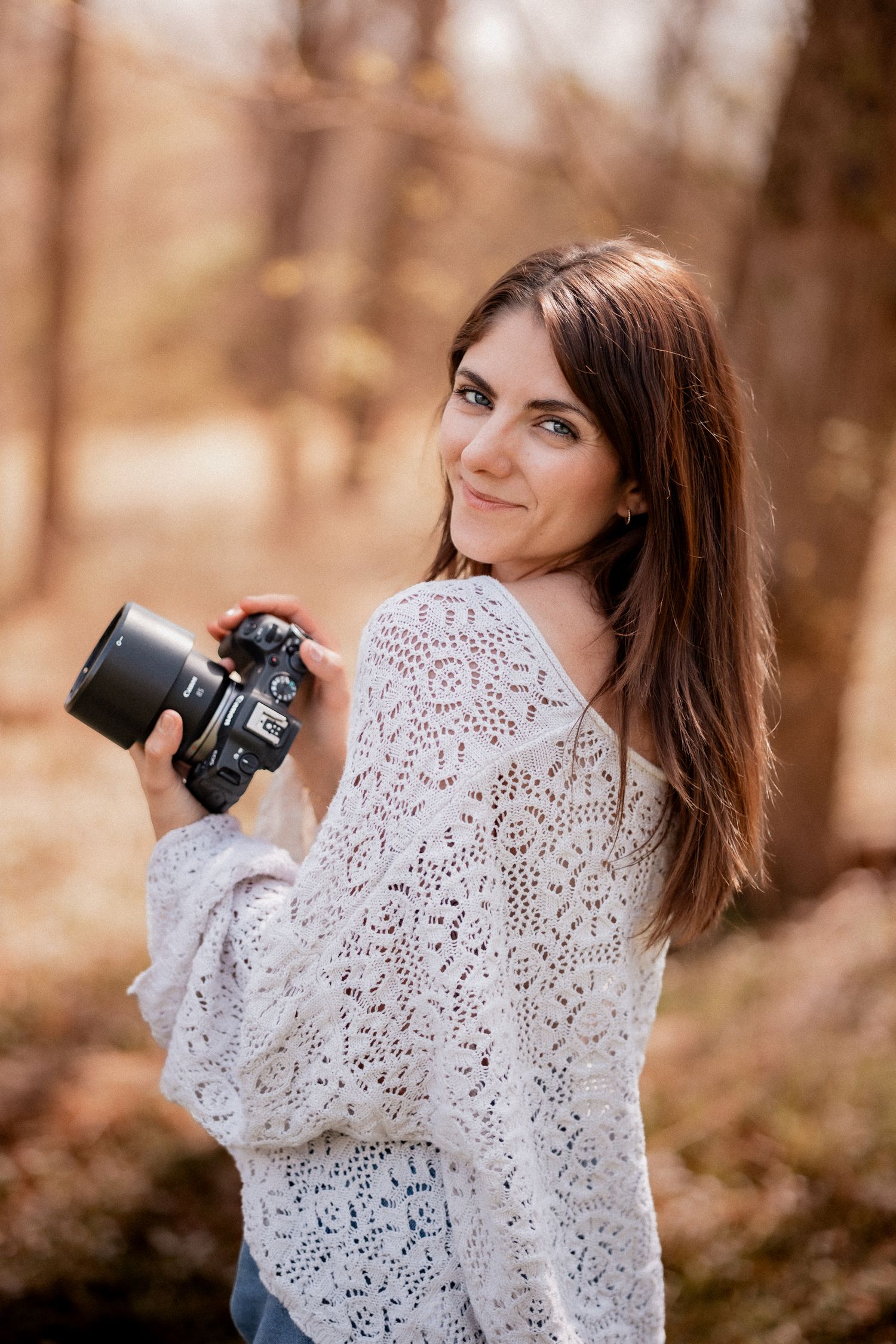 A smiling brunette woman holds a DSLR camera in an autumn forest, wearing a white lace knit sweater.