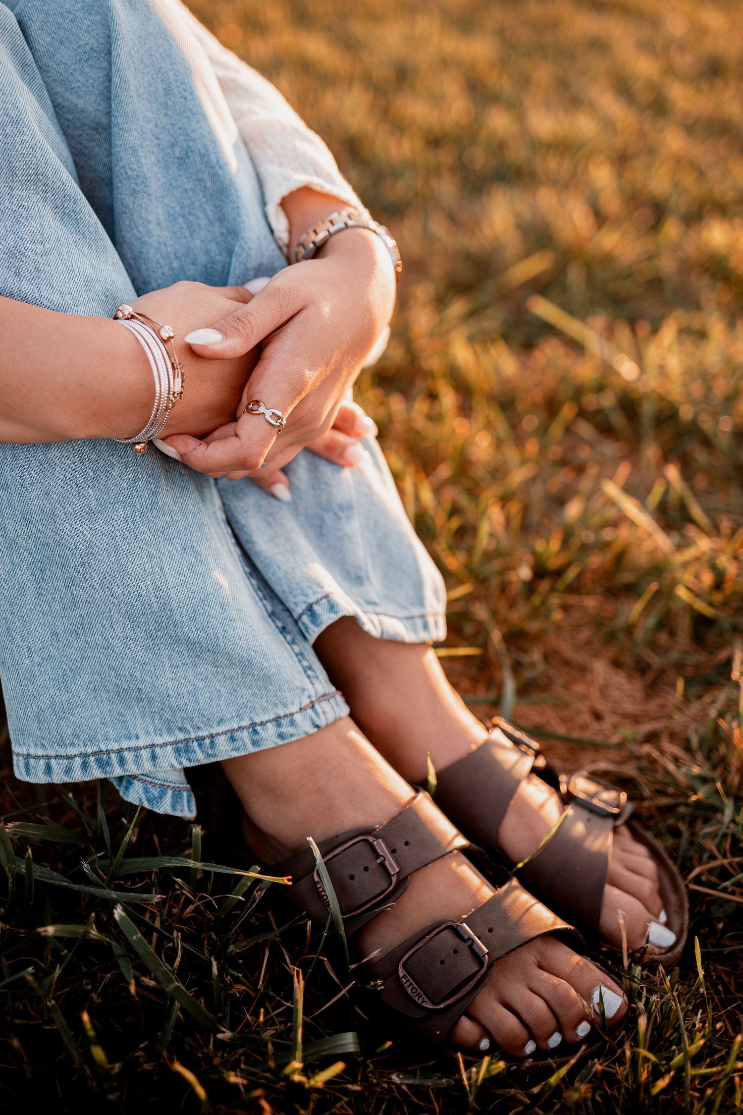 Close-up of brown sandals and jewelry on feet sitting in dry grass during sunset.