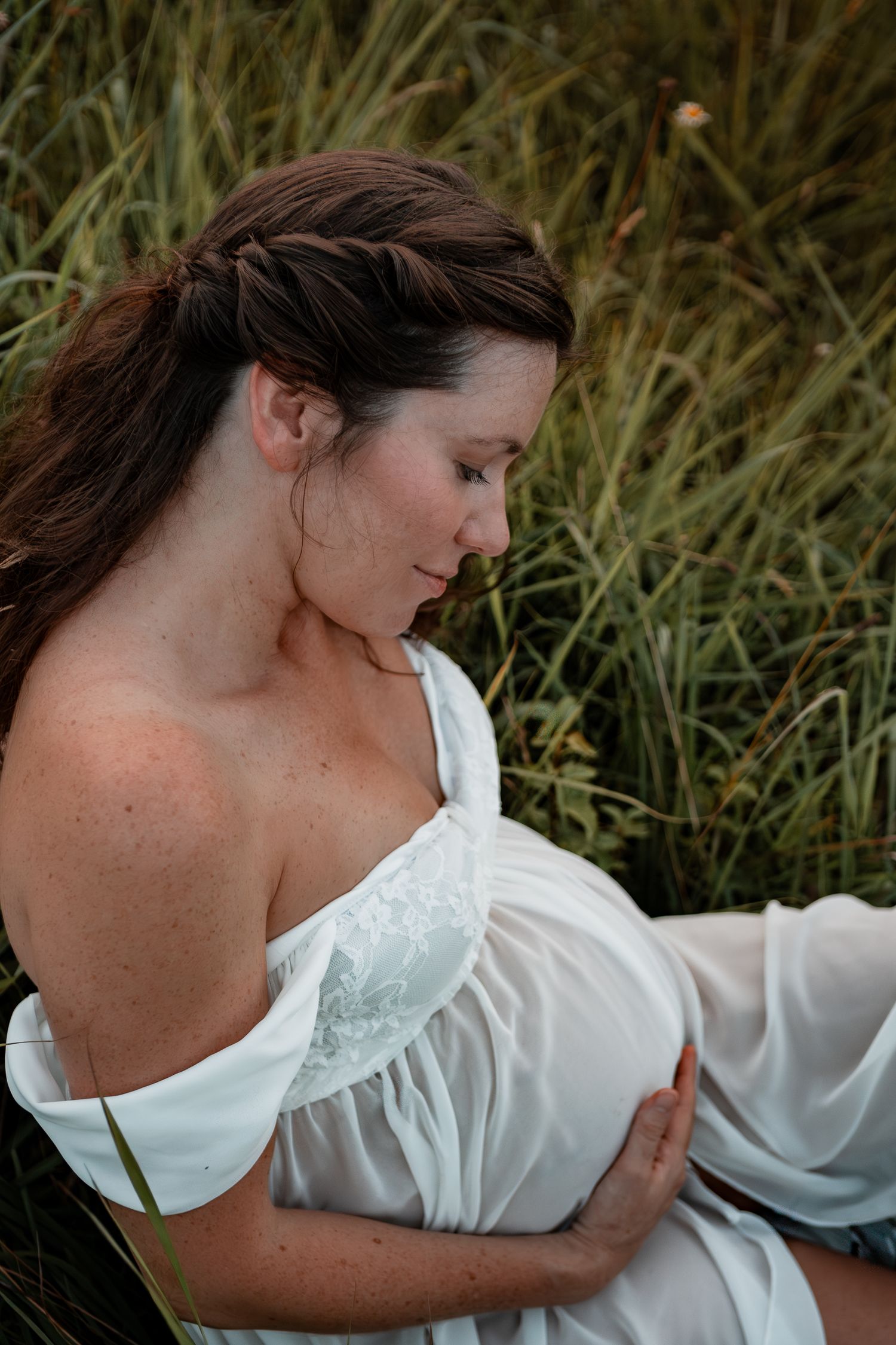 A woman in a white flowy dress relaxes thoughtfully in tall grass during an outdoor photo session.