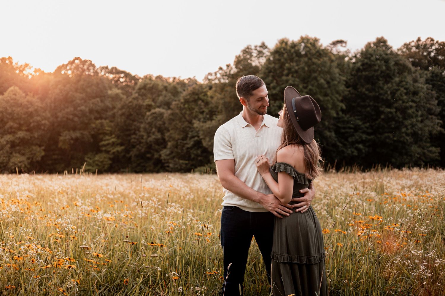 A couple embraces in a golden meadow at sunset with trees in the background.