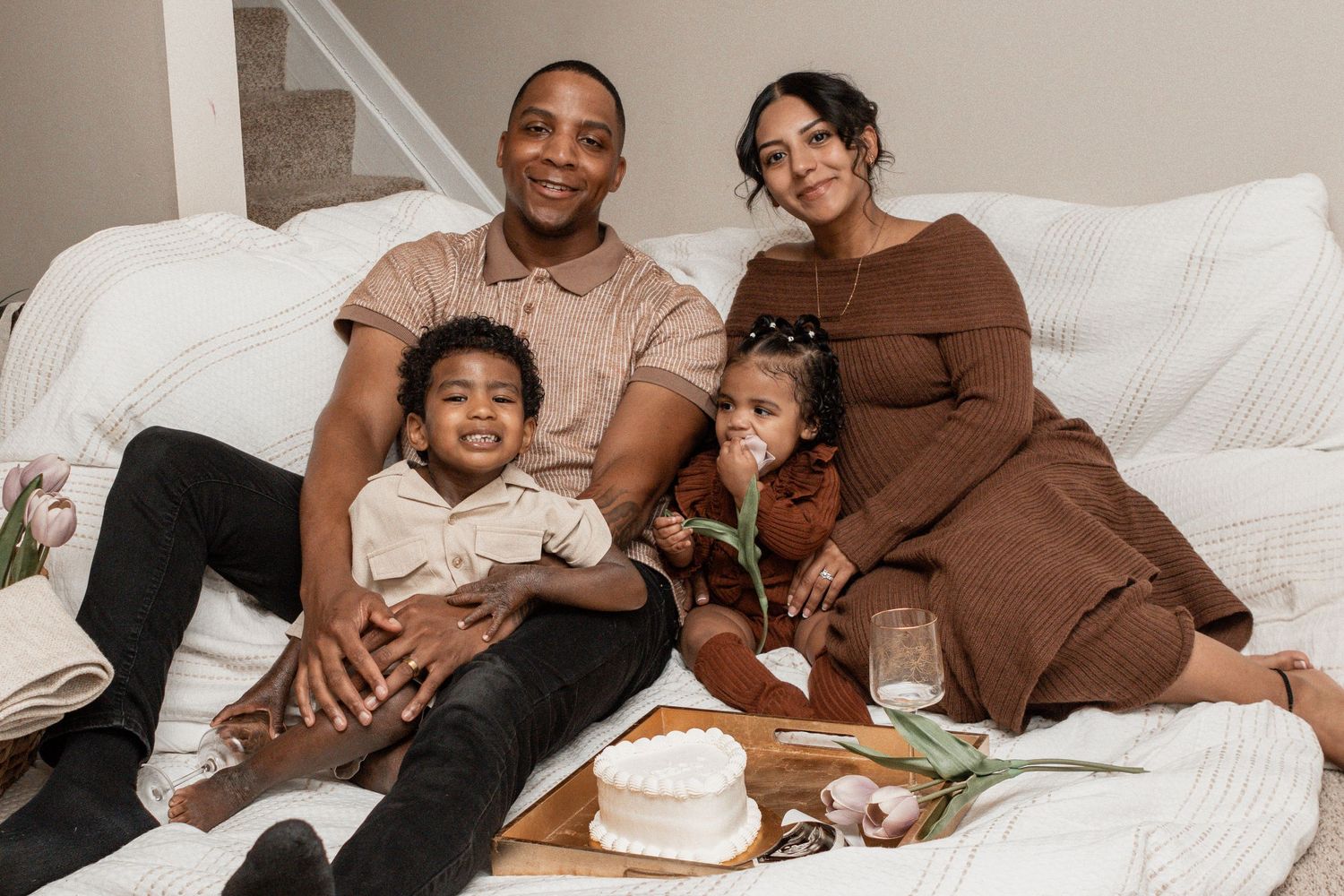 A family relaxes together on a white sofa with a cake and flowers at their feet.