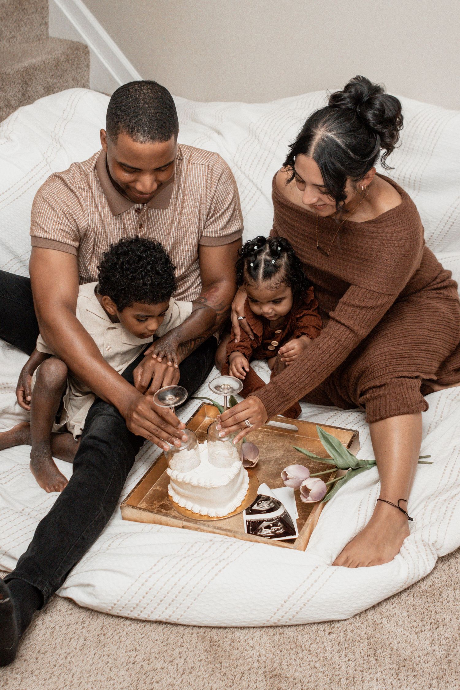 A family sits together on a white comforter looking at photos and a cake on a wooden board.