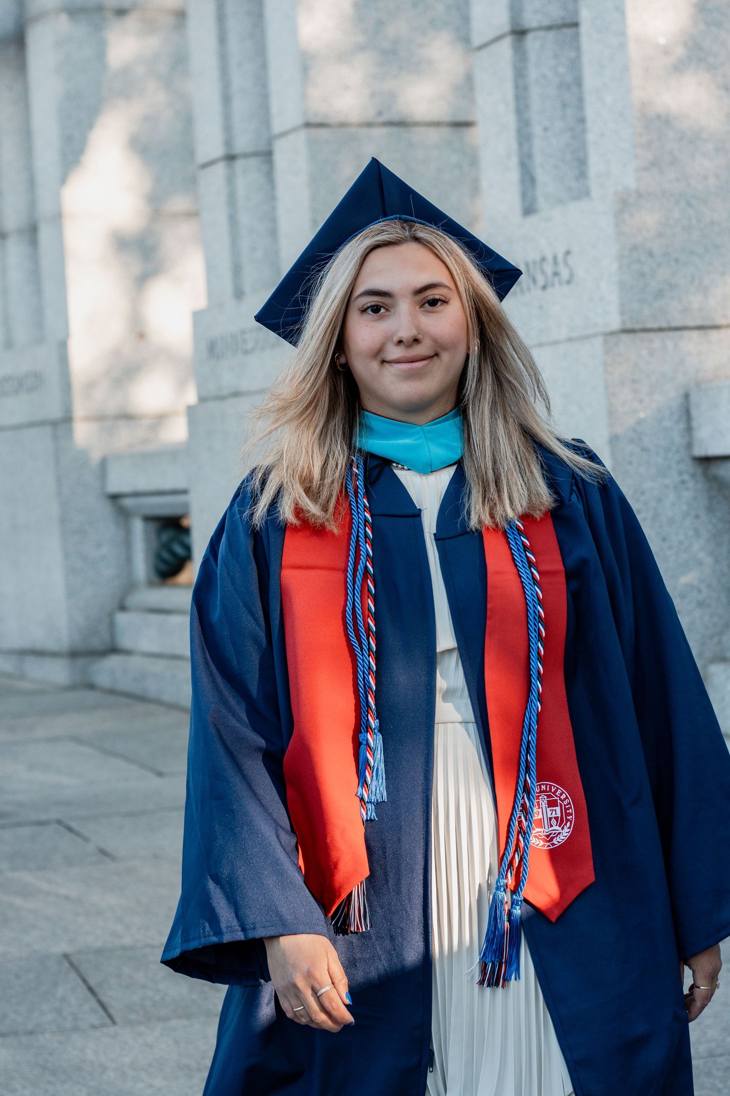 Graduate wearing cap, gown and honor cords poses beside stone columns during commencement.