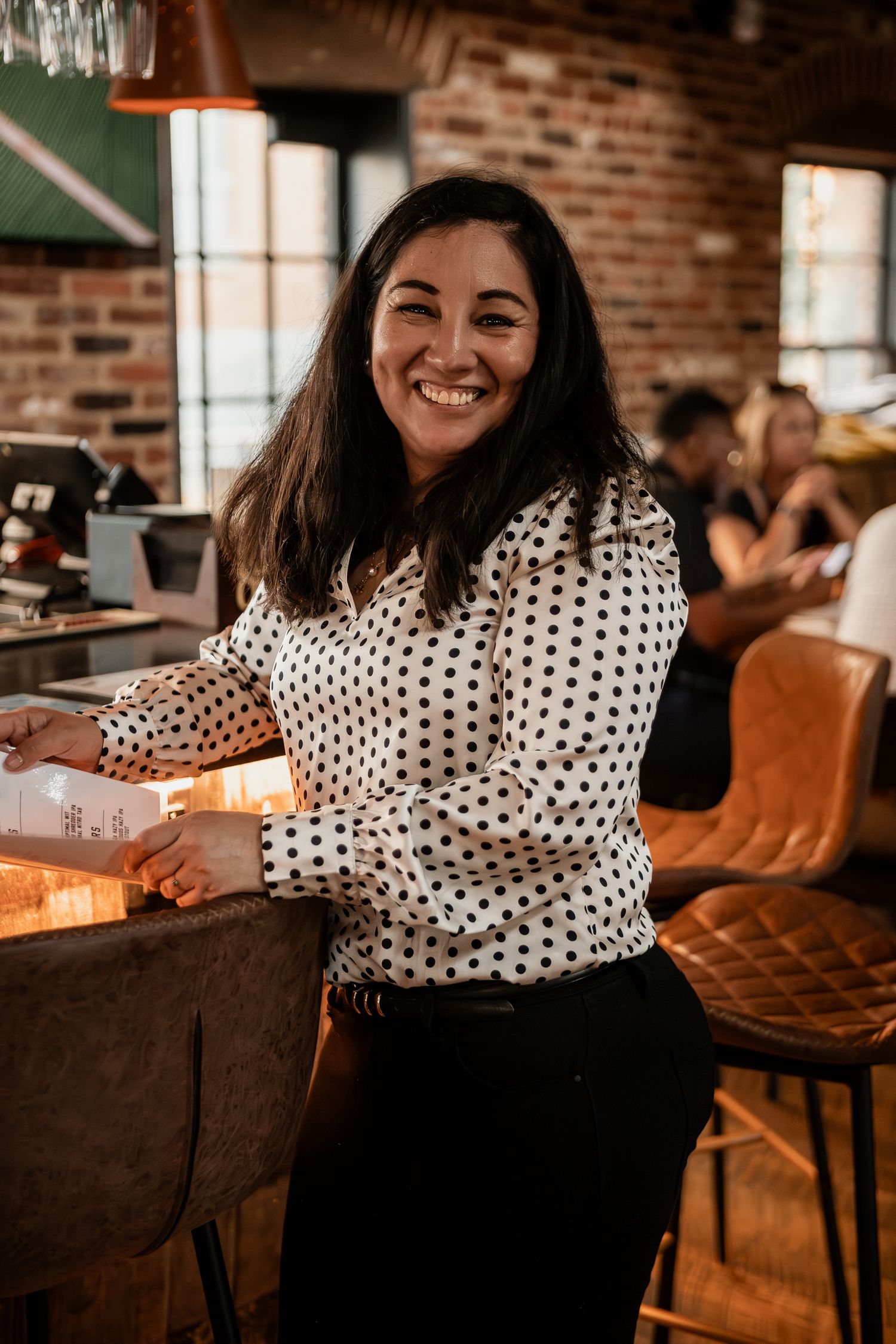 A person in a polka dot blouse poses at a bar with exposed brick walls and warm lighting.