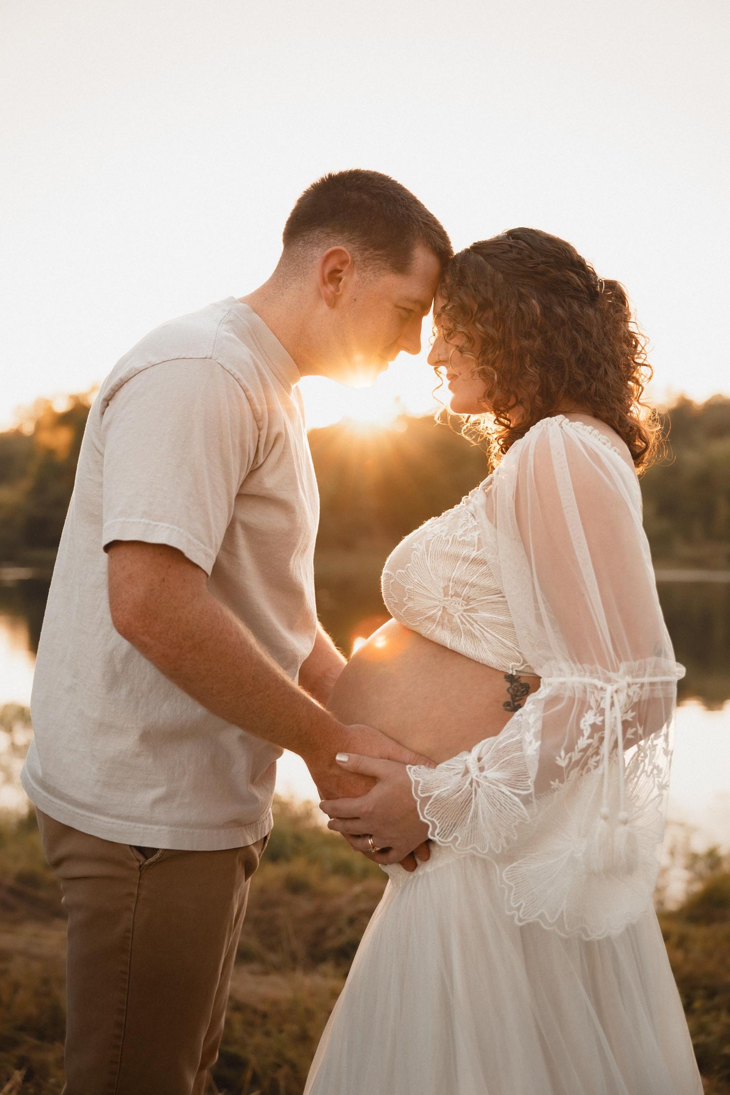 Couple holding pregnant belly at golden hour sunset by a lake, man and woman touching foreheads in tender maternity photo.