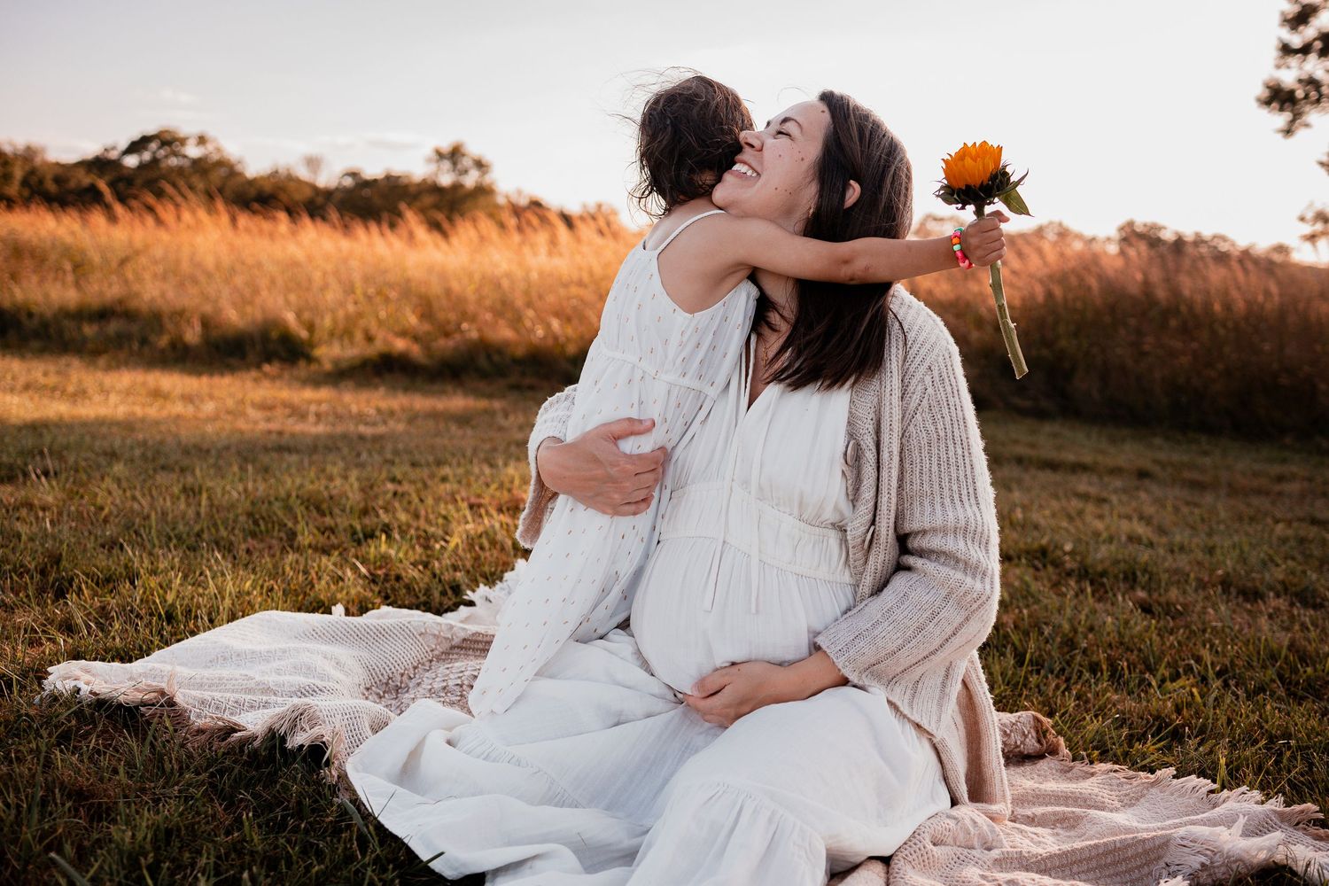 A romantic maternity photoshoot at sunset shows a couple embracing on a picnic blanket in a golden field.
