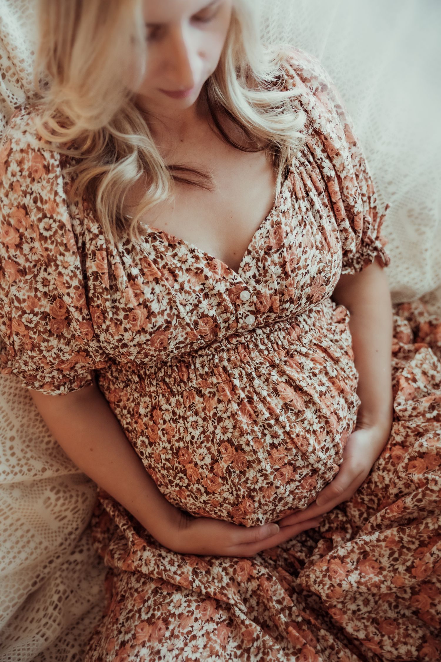 A pregnant woman in a floral dress sits on a cream blanket while gently cradling her baby bump.