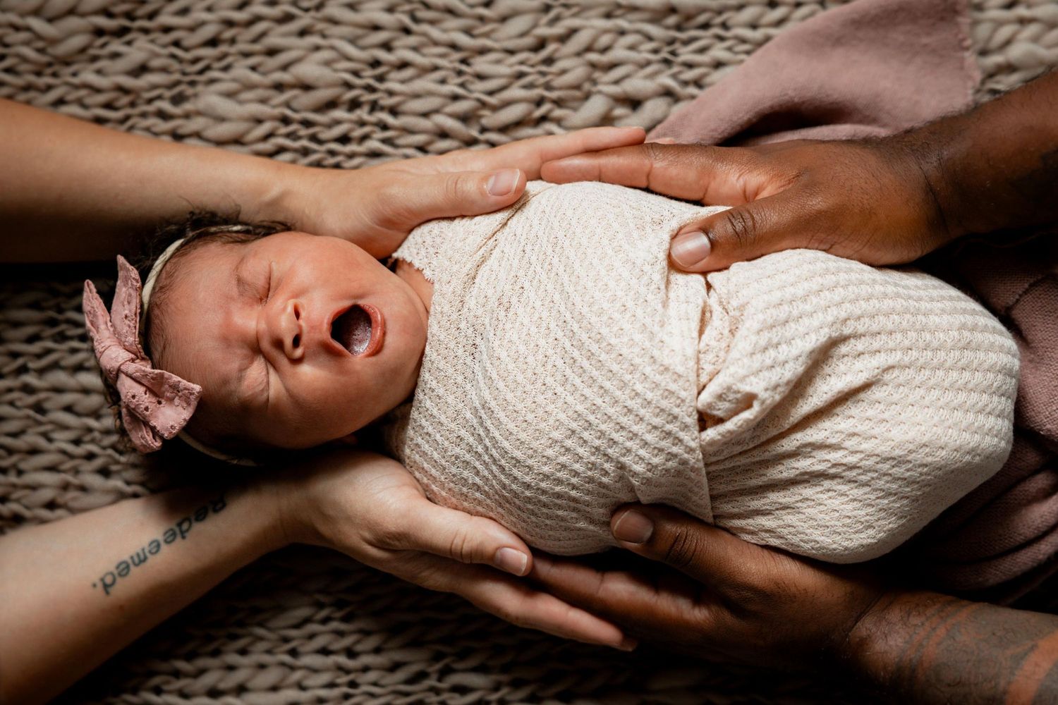 Newborn baby girl in cream swaddle with pink bow headband, yawning while parents gently cradle her.