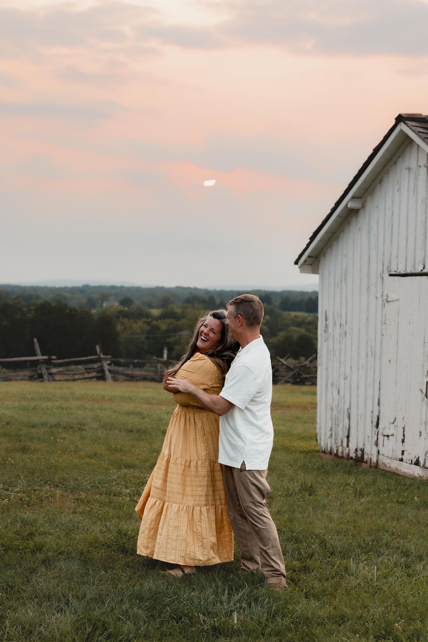 A laughing couple embraces near a white barn at sunset during a golden hour outdoor photo session.