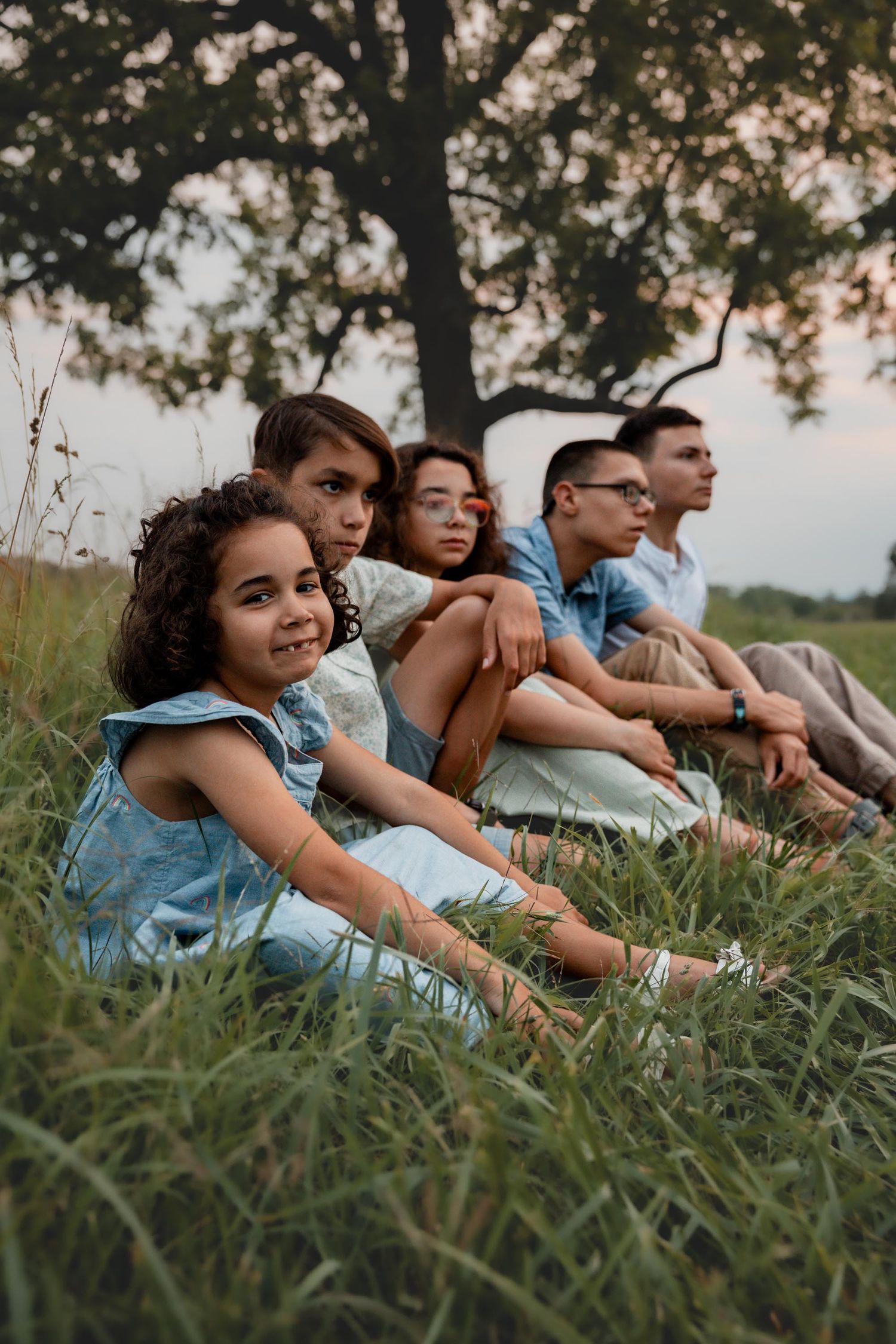 Four siblings sit together in tall grass beneath a large tree during a relaxed outdoor family session.