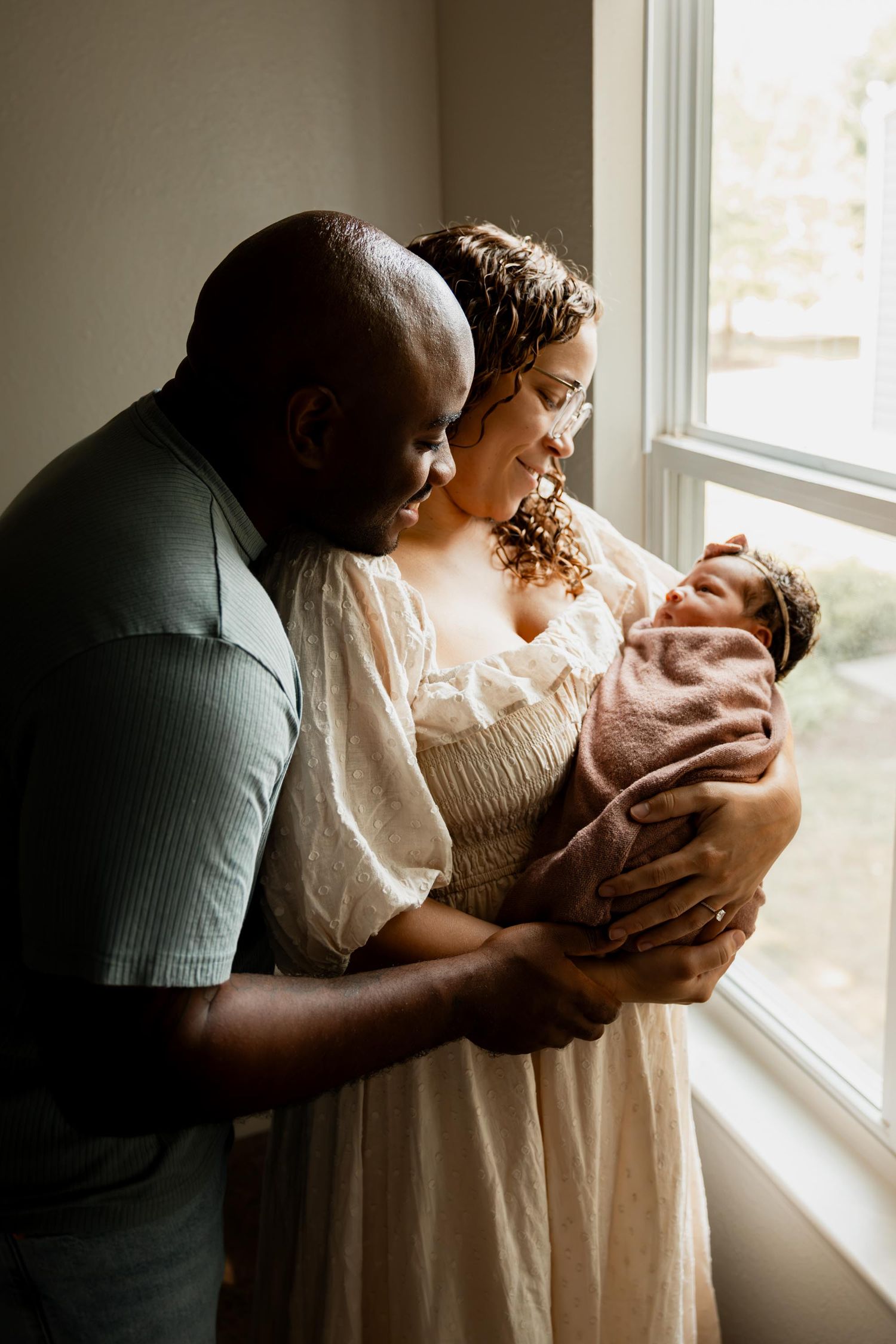 A couple lovingly gazes at their newborn baby wrapped in pink by a sunlit window.