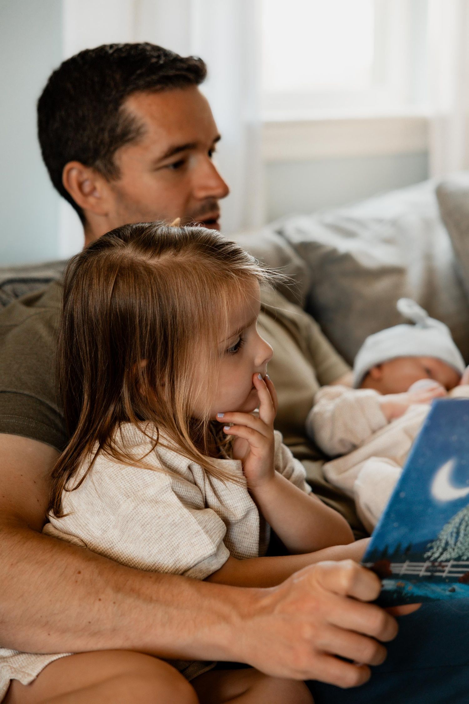 Father reads a picture book to toddler daughter while holding newborn baby on couch at home.