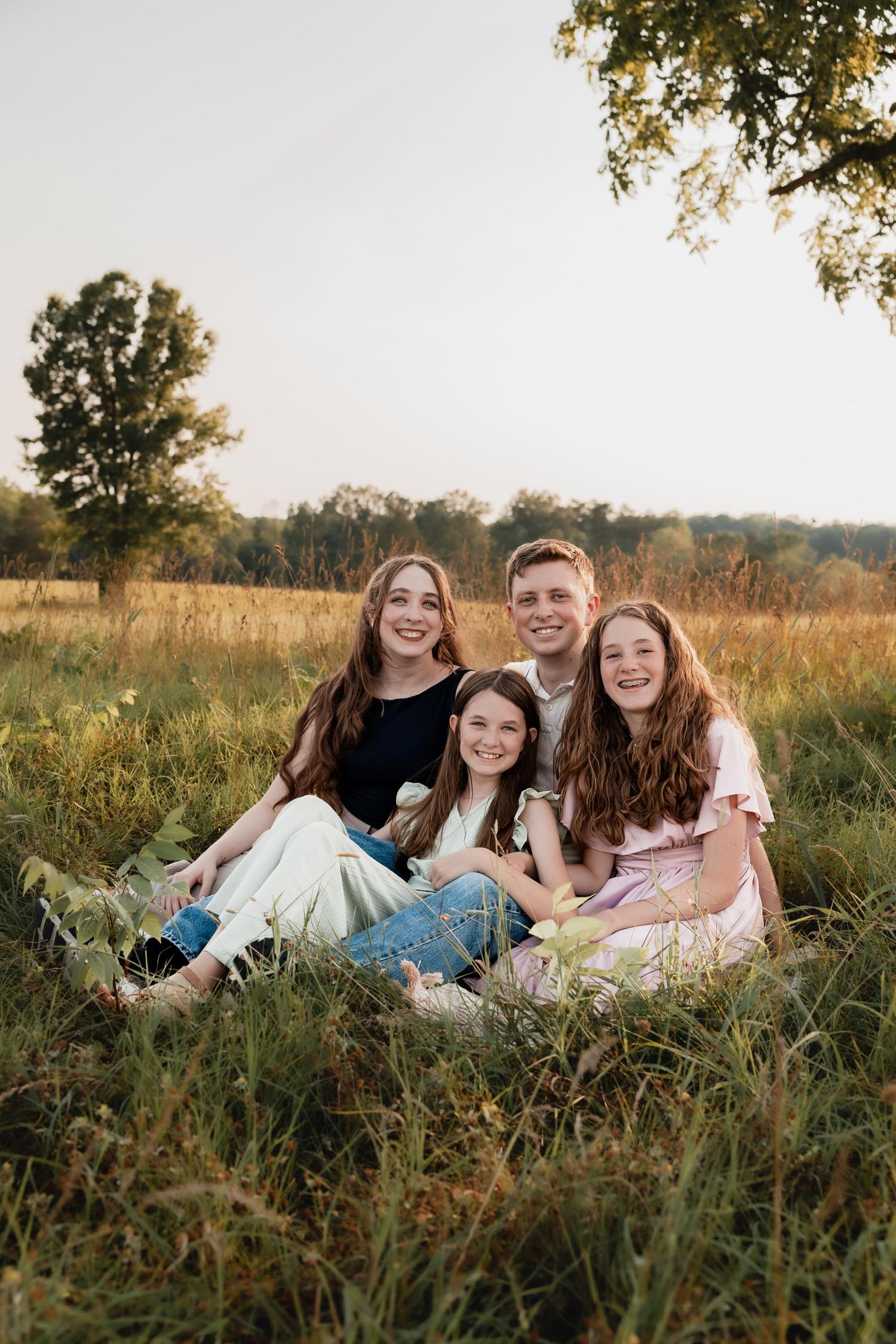 Four siblings sitting together smiling in a golden field during a sunny outdoor family photo session.