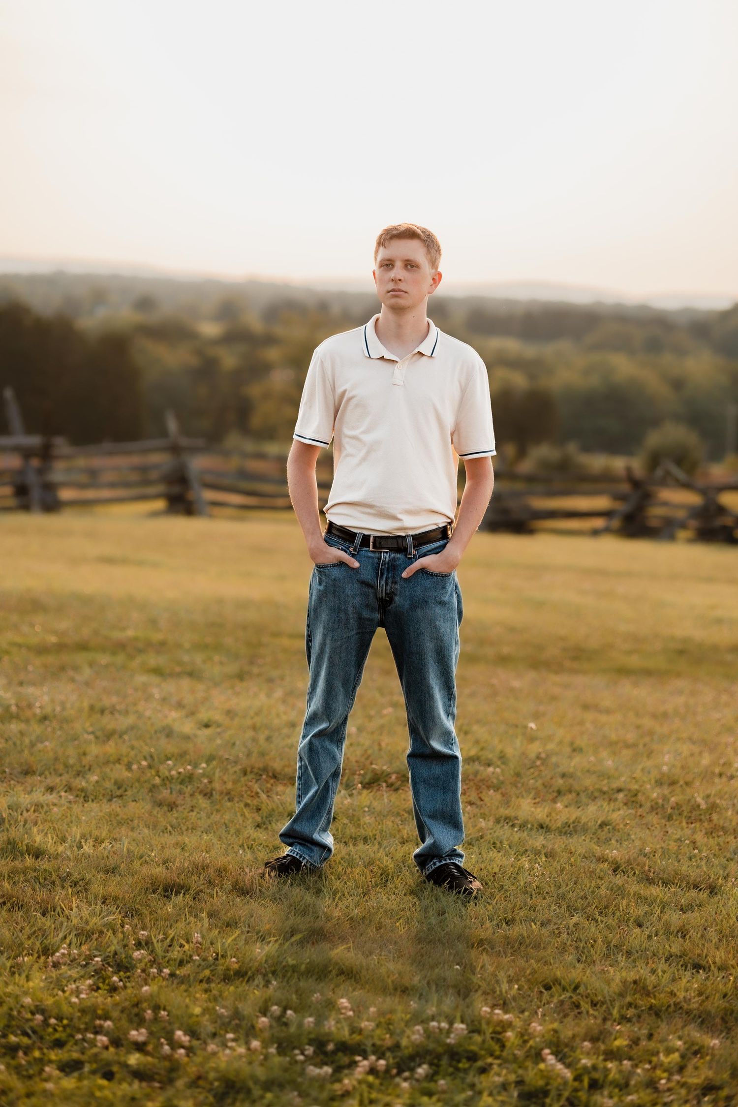Young man in white polo shirt and jeans stands confidently in a scenic field at golden hour.
