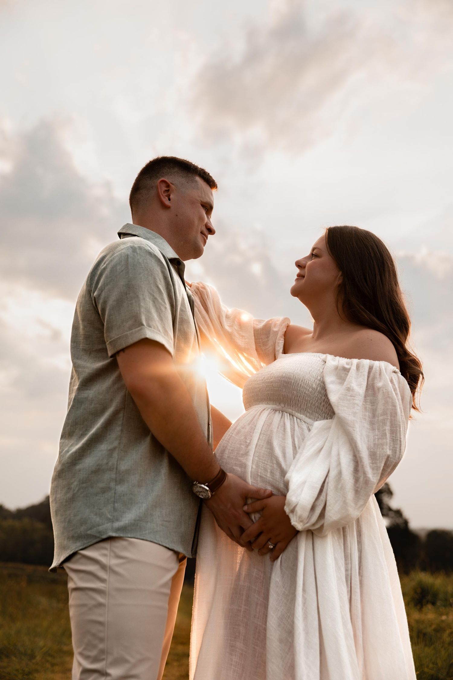 A couple shares a tender moment at sunset, the woman in a white maternity dress, holding hands in a golden field.