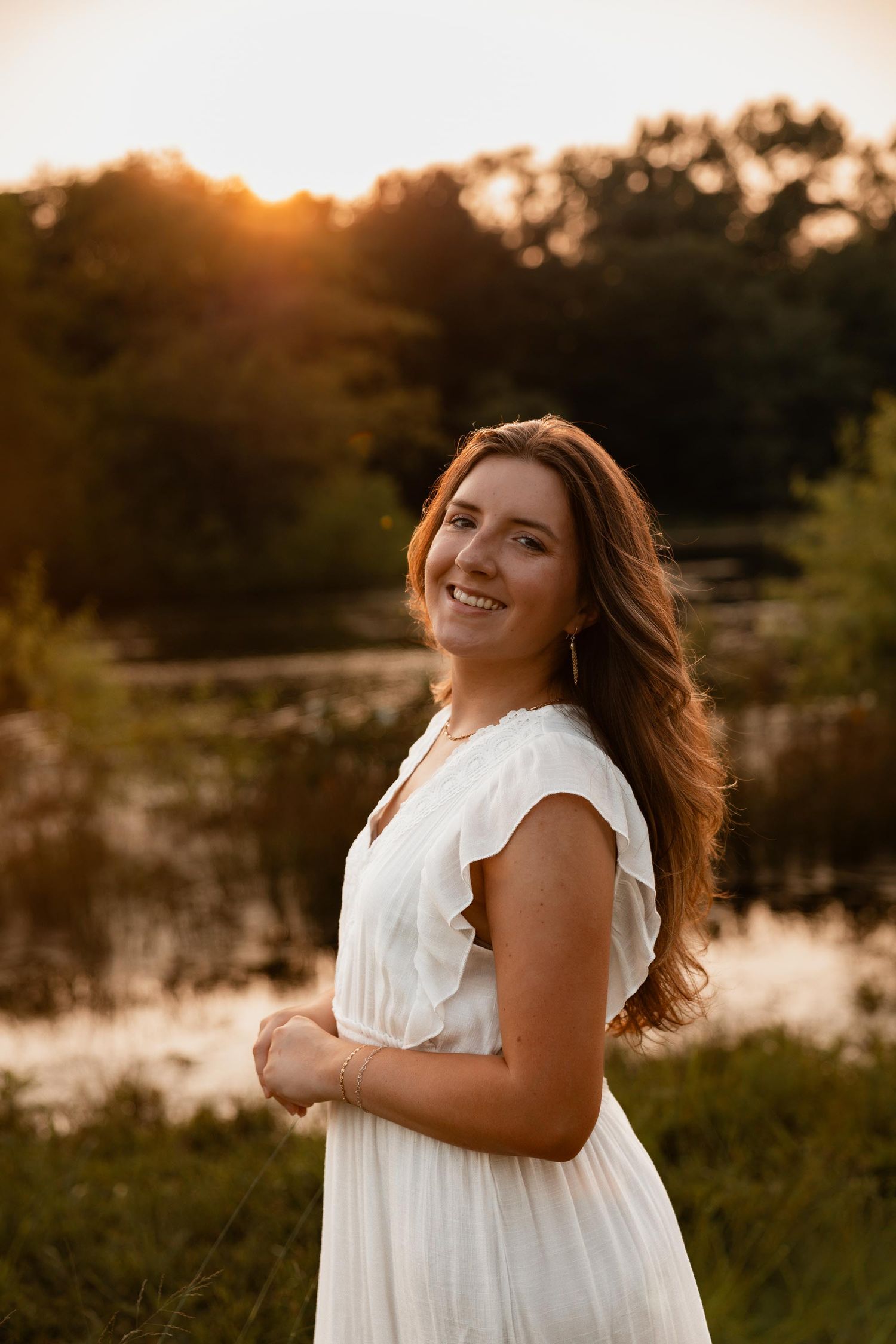 Smiling woman in white dress at golden hour sunset near a serene lake surrounded by lush green trees.