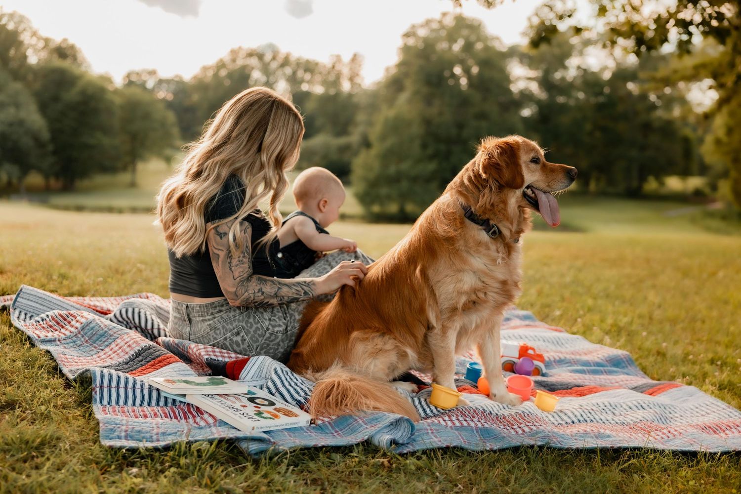 Woman with baby and golden retriever dog enjoying a picnic on a colorful quilt blanket in a sunny park.