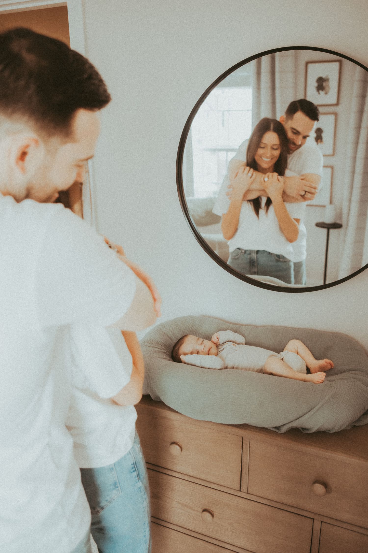 A tender family moment captured in a circular mirror reflection above a dresser showing parents with their baby.