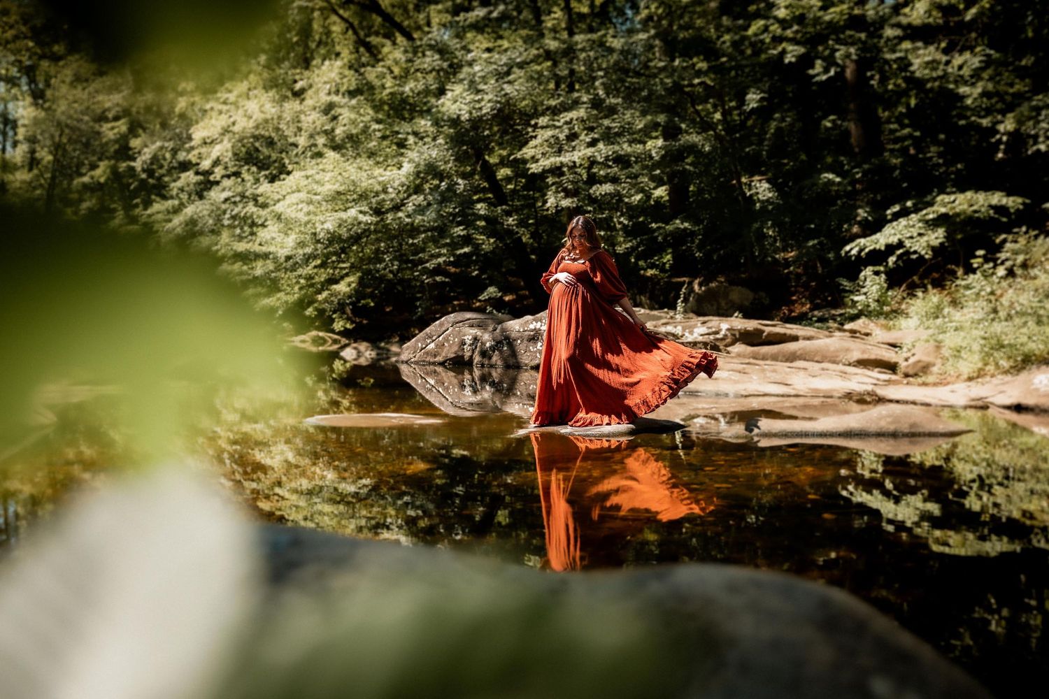 Person in flowing red dress standing on rocks beside a reflective forest river surrounded by lush green trees.