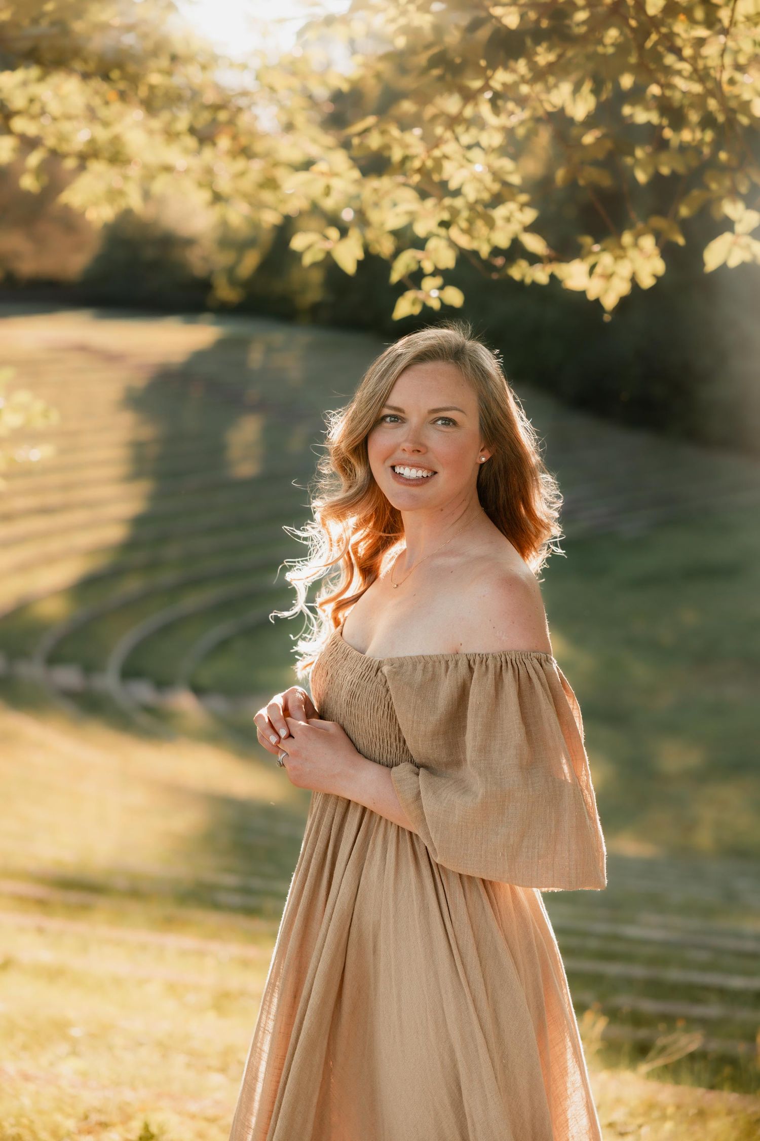 Smiling woman in golden off-shoulder dress standing in sunlit park with terraced lawn background.