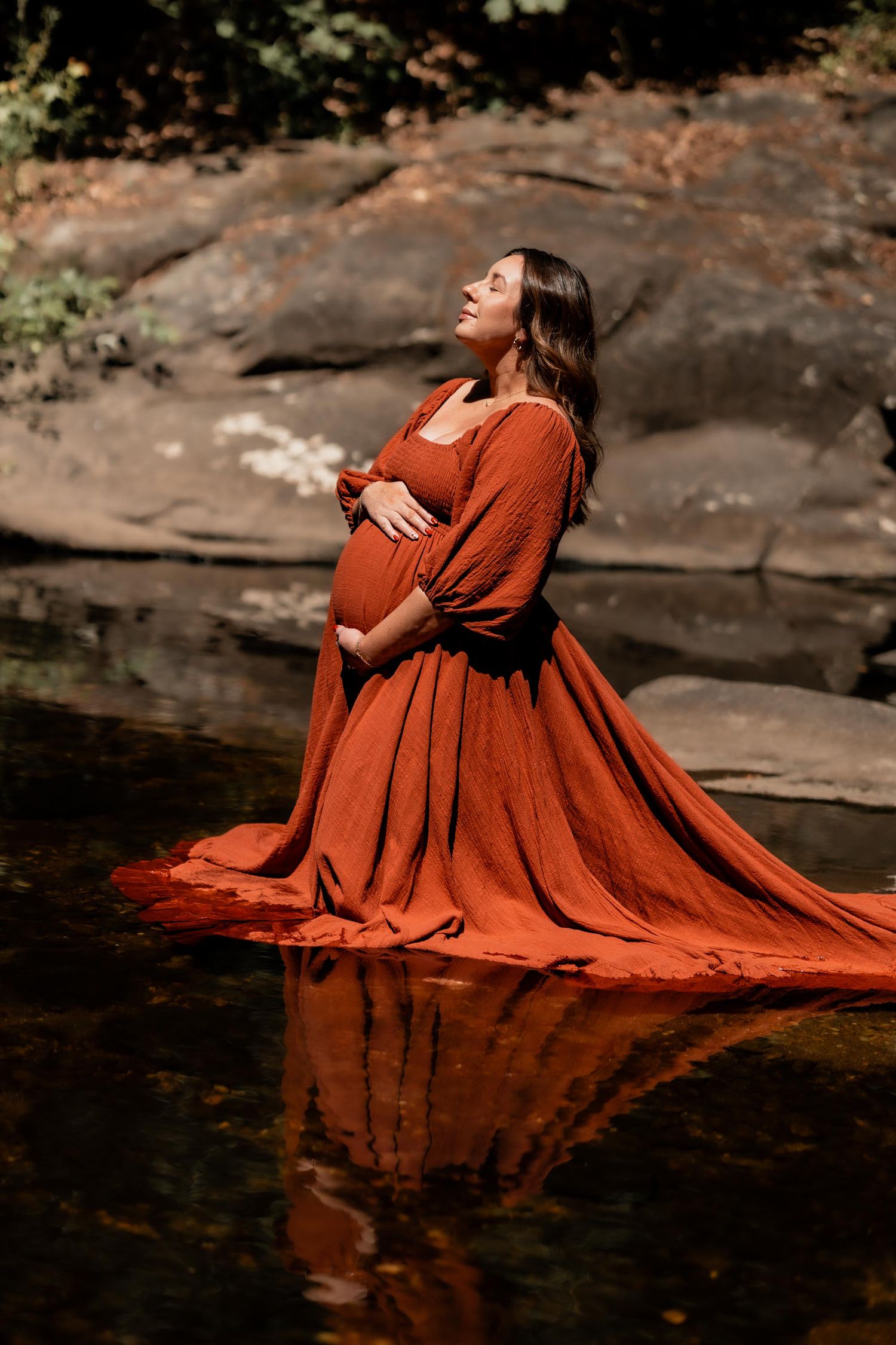 Pregnant woman in rust orange gown standing in shallow creek, cradling belly, surrounded by rocks and foliage.