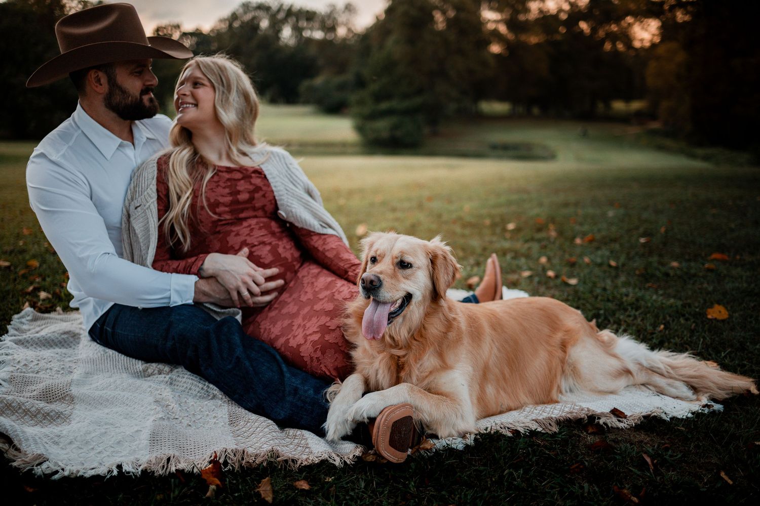 A couple sits on grass with their golden retriever during a sunset maternity photo session in a park.