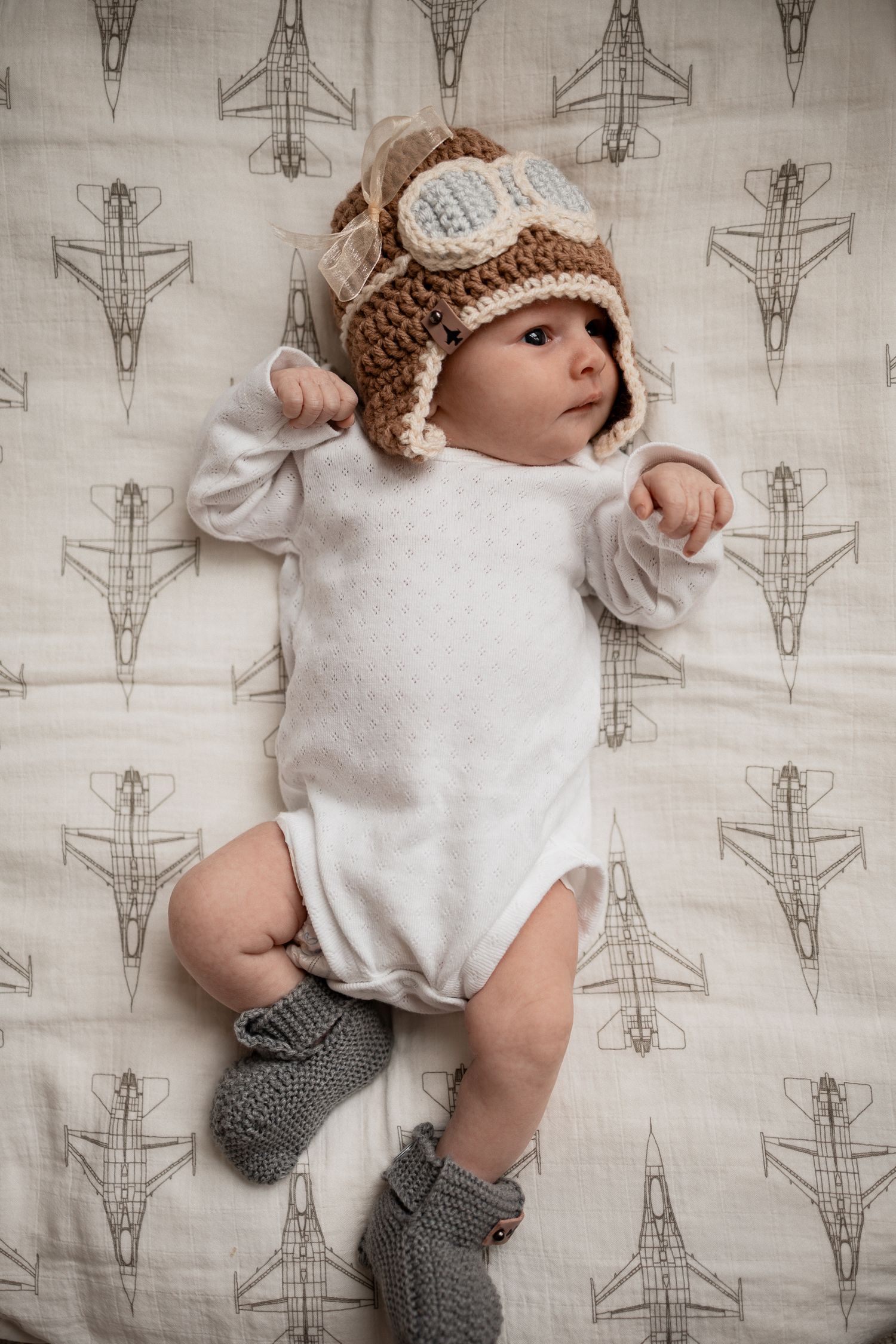 Baby wearing white onesie and knit hat lies on airplane patterned fabric with gray knit booties.