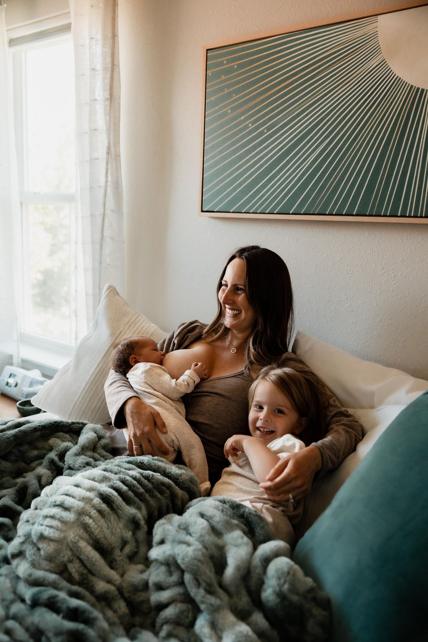 Smiling mother sitting on bed with newborn baby and toddler daughter, wrapped in teal chunky knit blanket.