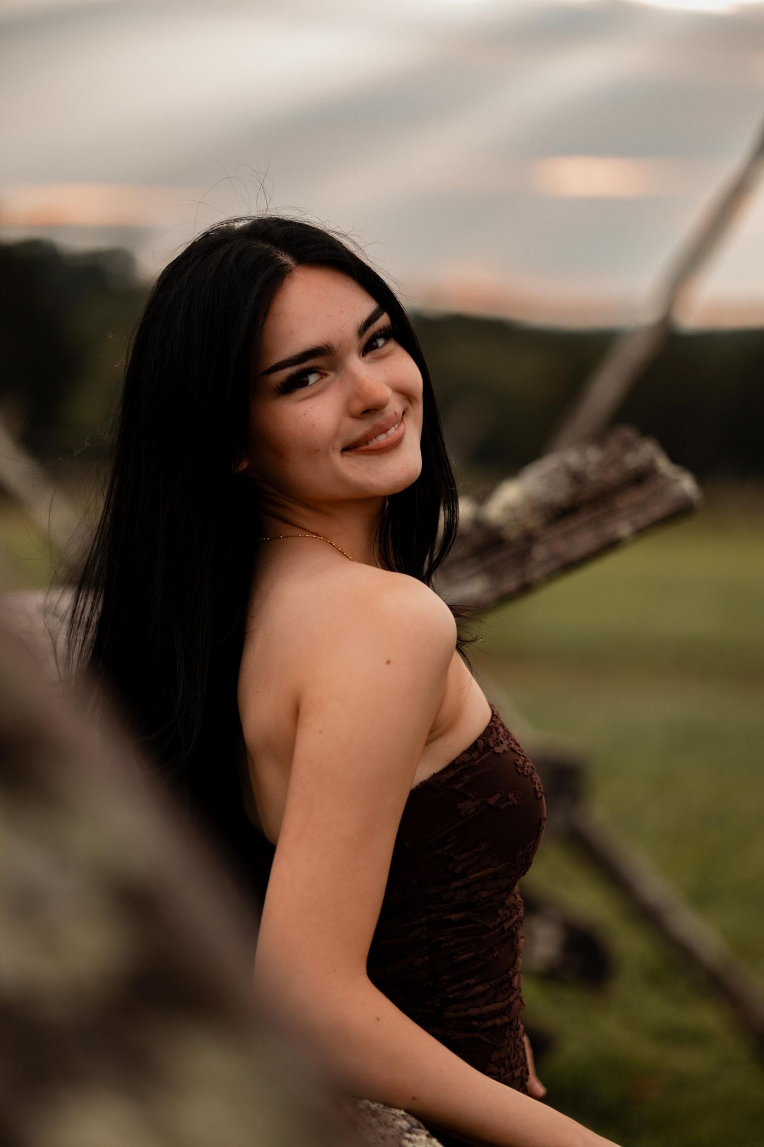 Young woman with long black hair smiling outdoors in a brown strapless dress against a scenic dusk sky.