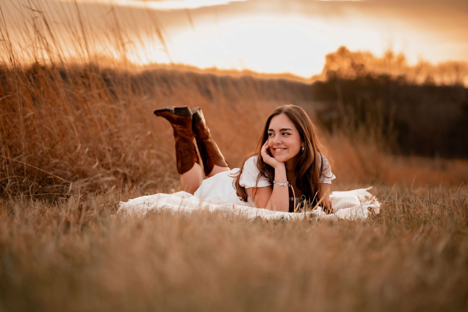 Young woman in white dress and brown boots lying in golden field at sunset, smiling dreamily.