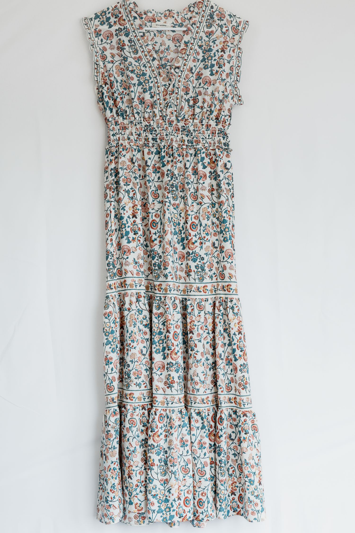 Floral maxi dress with blue and brown pattern featuring tiered skirt and elastic waist hanging against white wall.