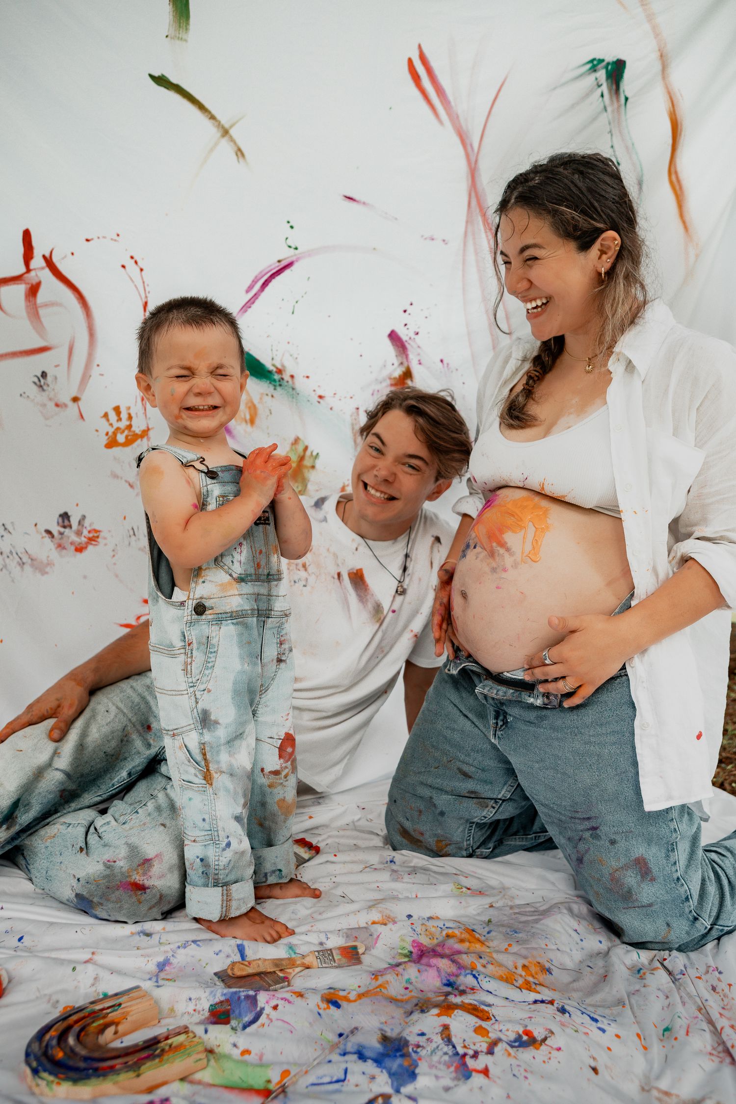 Joyful family having fun with colorful paint splatters on a white backdrop during creative art session.