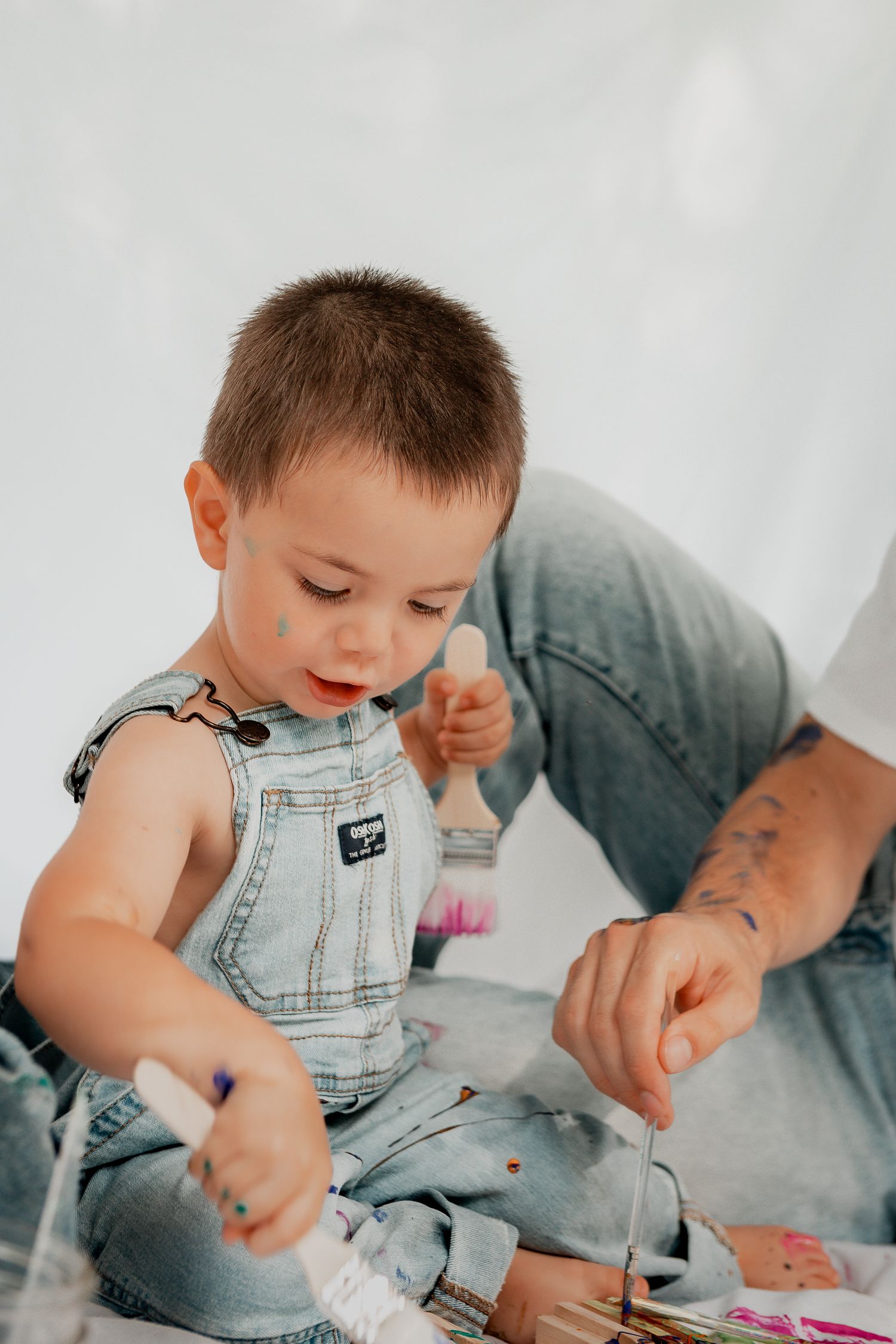 A small child in denim overalls sits on the floor engaged in play while an adult hand points nearby.