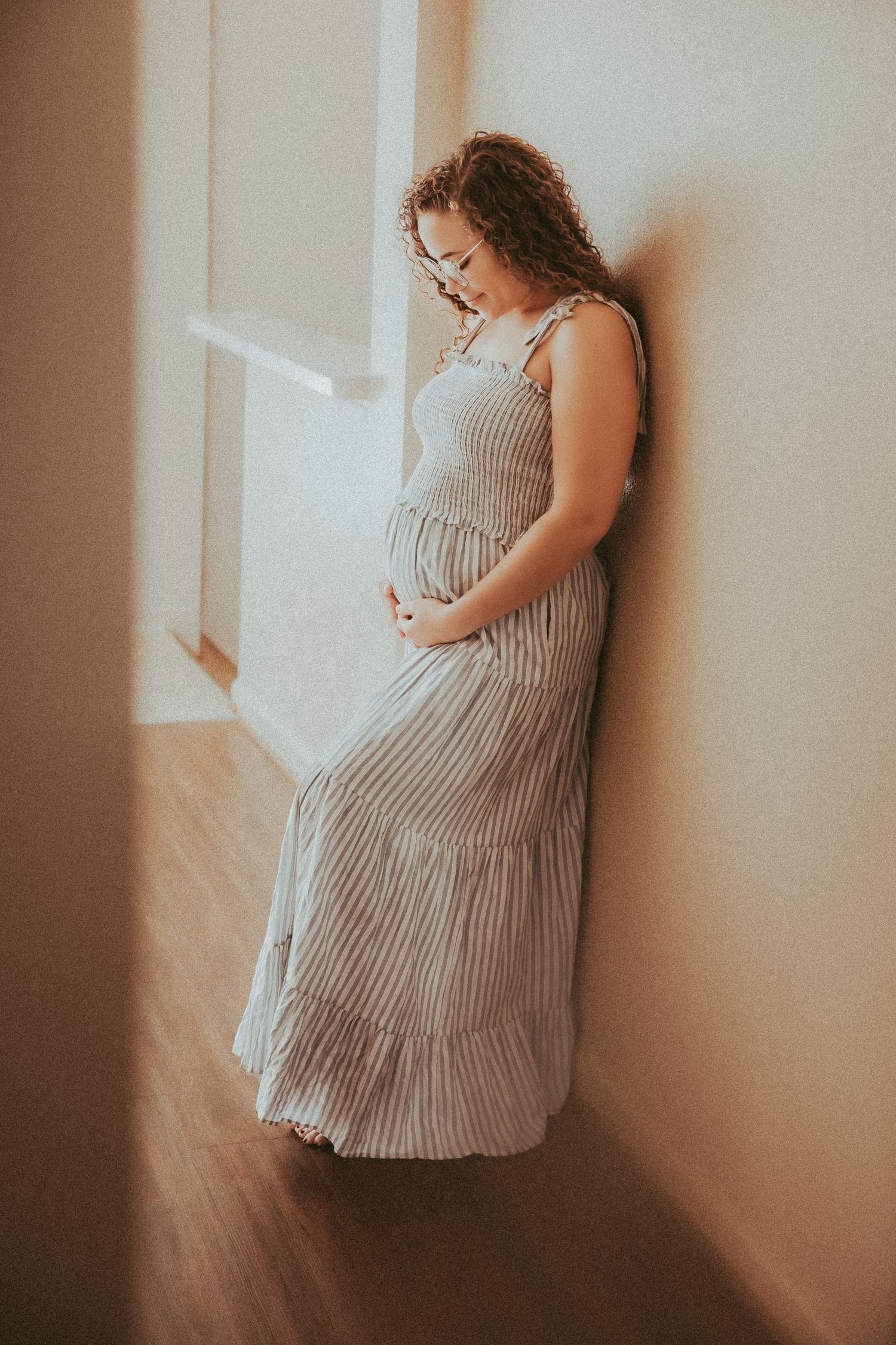 A pregnant woman in a striped dress stands near a window in soft natural lighting creating a serene maternity portrait.