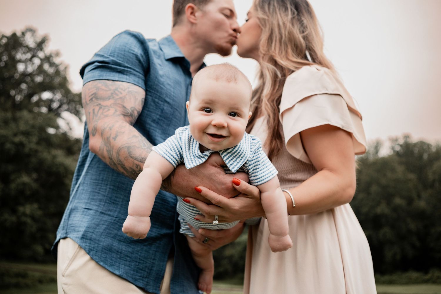 A happy baby in a blue outfit smiles while being held between two parents embracing outdoors.